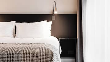 Une chambre d’hôtel moderne avec un grand lit, une table de chevet avec une lampe et une vue sur l’extérieur à travers des rideaux légers.
