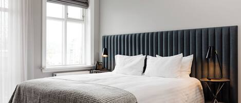 Una habitación de hotel moderna con una cama grande, un cabecero oscuro, una mesita de noche con lámpara y una ventana con cortinas.