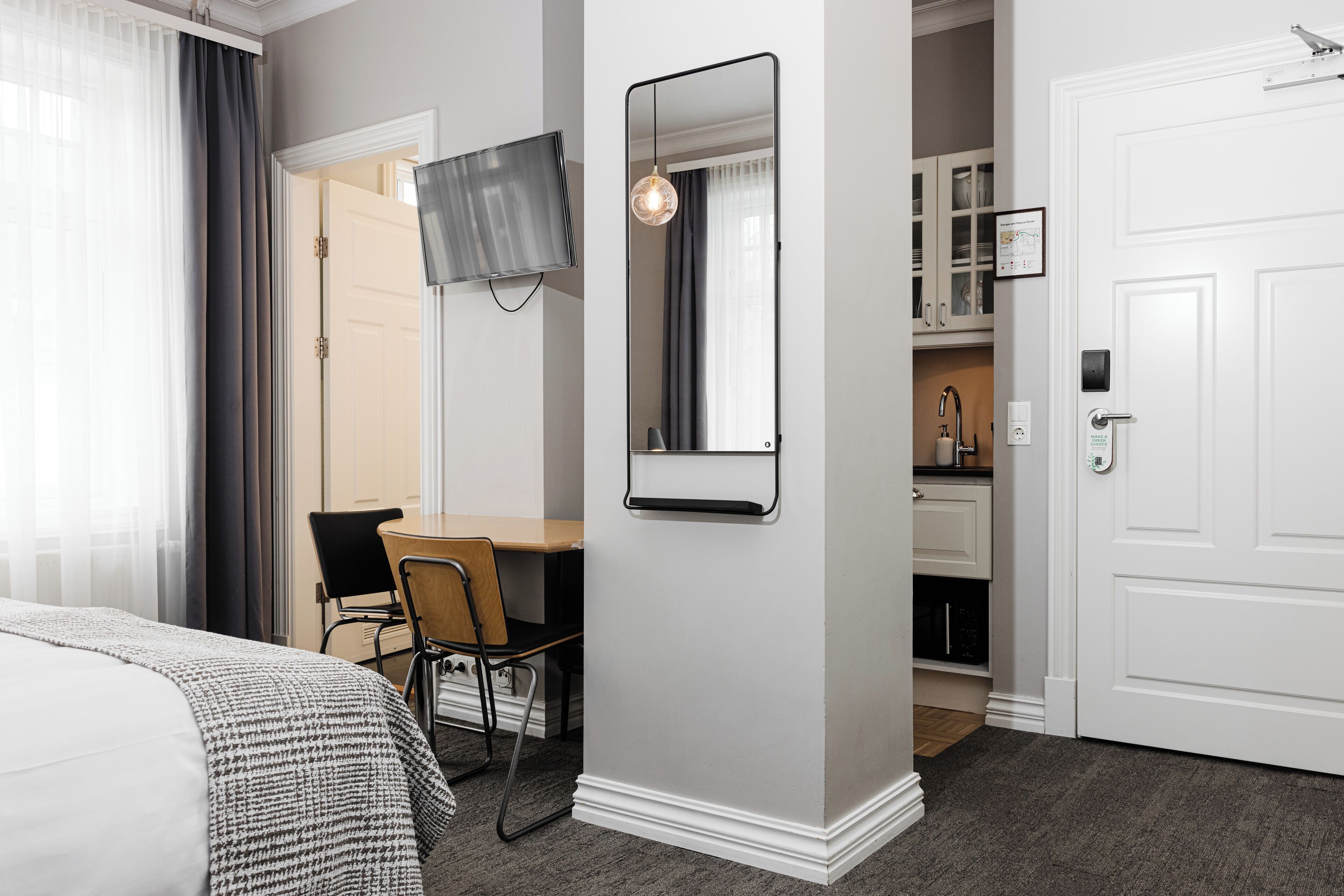 A hotel room with a bed, a desk, a chair, a TV, a mirror, a door, and a small kitchenette.