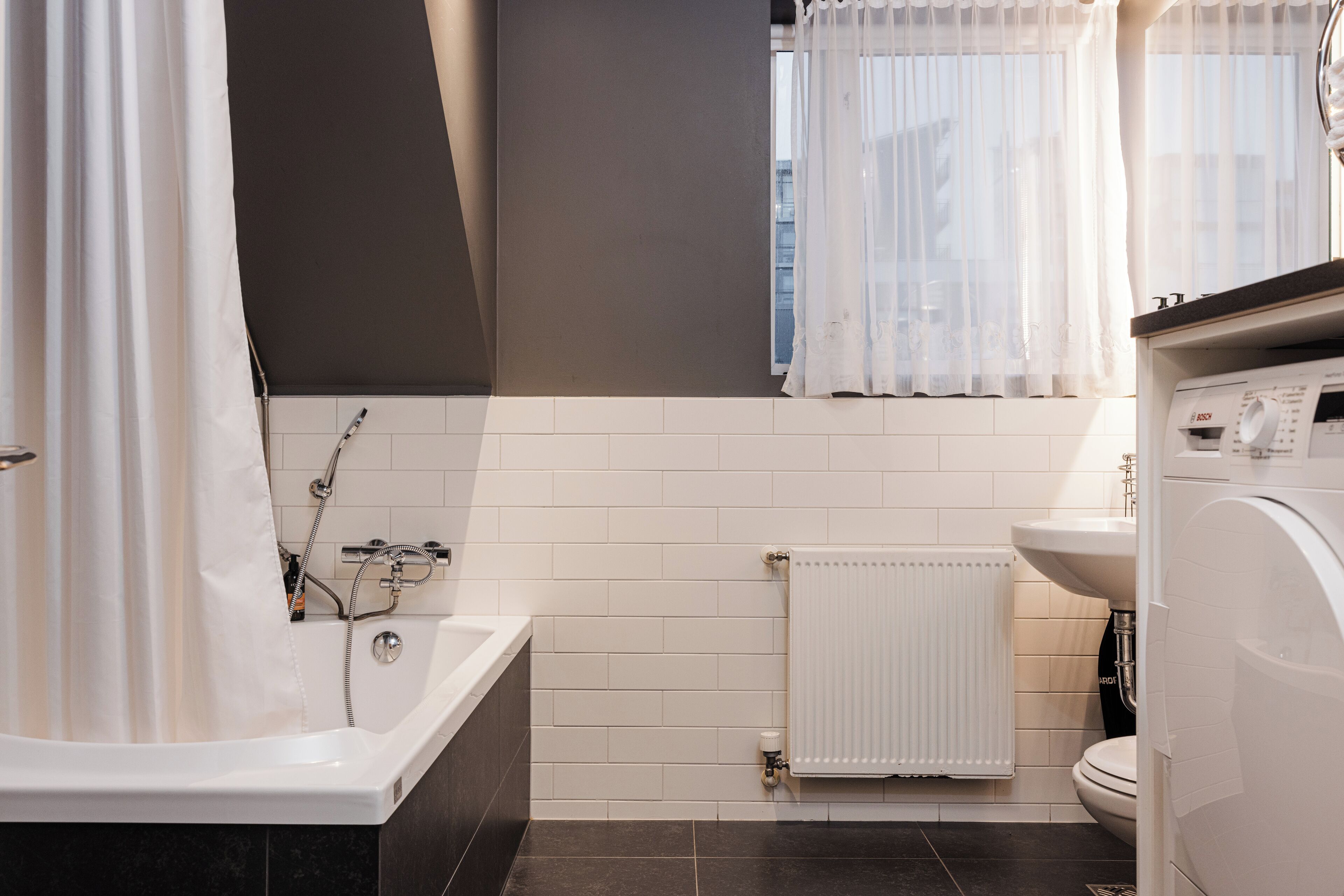 A modern bathroom with a bathtub, a toilet, a sink, and a window with sheer curtains.