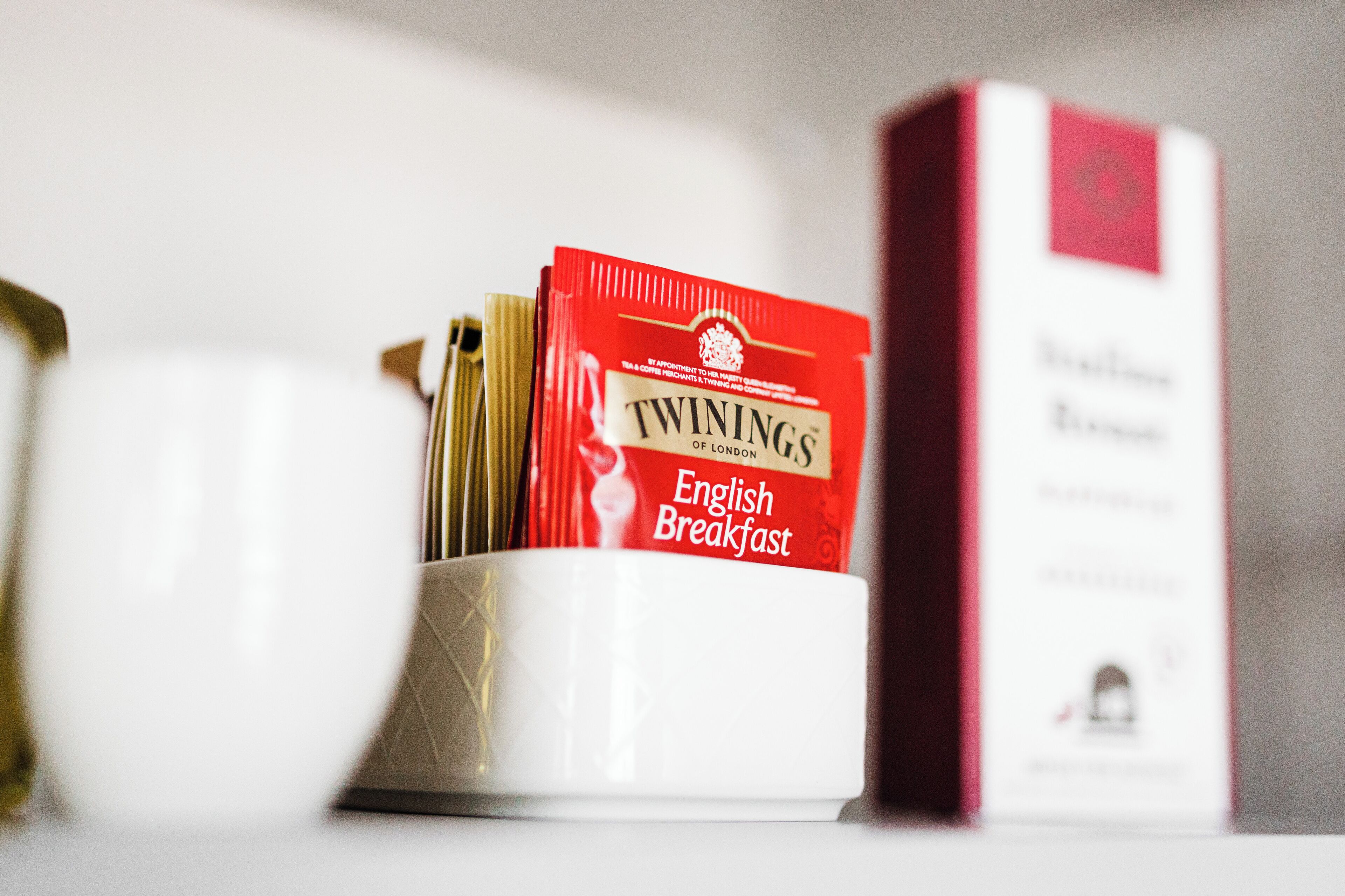 Una taza blanca con un diseño dorado, que contiene un sobre rojo con la etiqueta "TWININGS English Breakfast".
