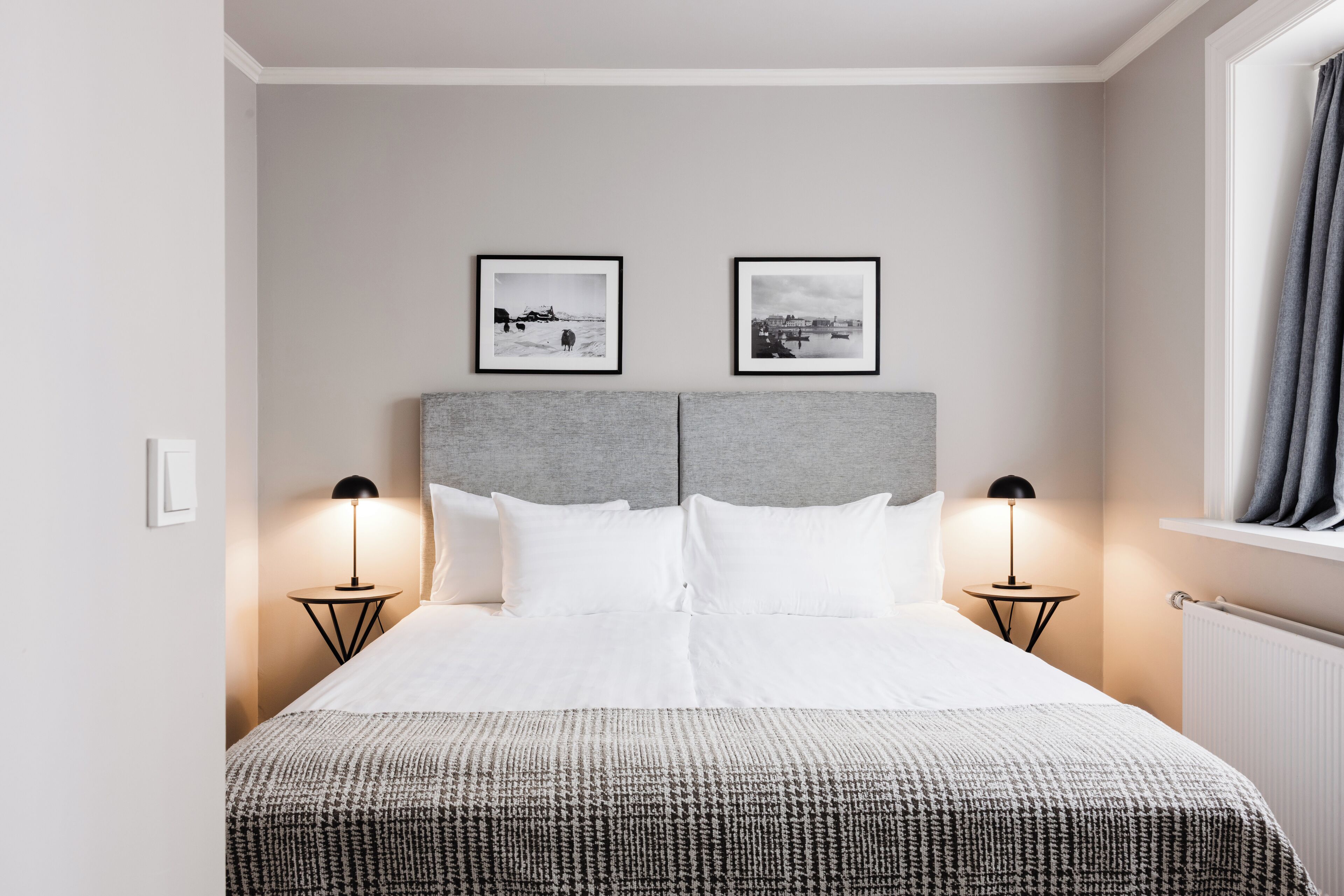 Cama bem arrumada com lençóis brancos e manta estampada, com duas mesas de cabeceira e abajures de cada lado, num quarto com cabeceira cinza e duas obras de arte emolduradas a preto sobre a cama.