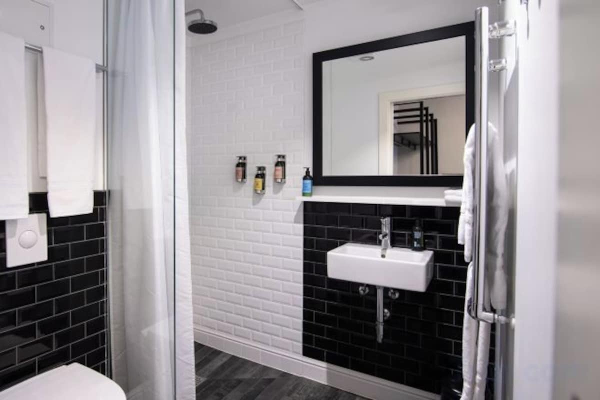 Une salle de bain avec un lavabo blanc, des murs recouverts de carreaux noirs, un miroir et un espace douche.