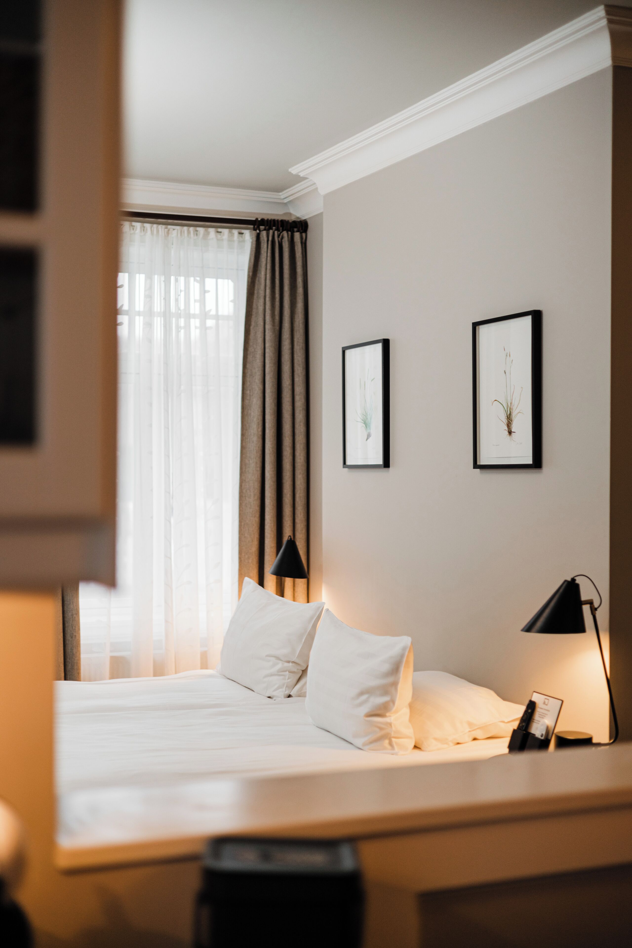 A hotel room with a bed, two framed pictures on the wall, a nightstand with a lamp, and a window with curtains.
