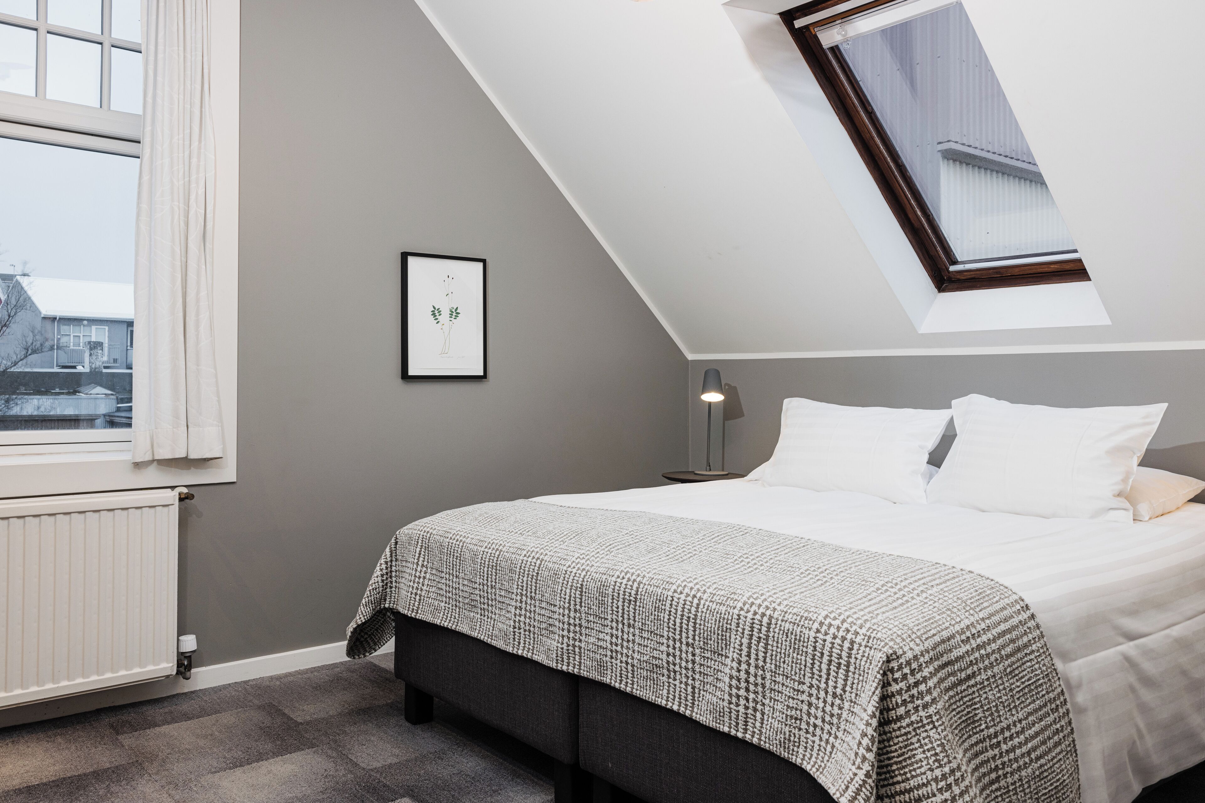 Un dormitorio con techo inclinado, una cama con ropa de cama blanca, una pared gris, un cuadro enmarcado y una ventana con cortinas.