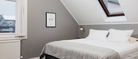Un dormitorio con techo inclinado, una cama con ropa de cama blanca, una pared gris, un cuadro enmarcado y una ventana con cortinas.