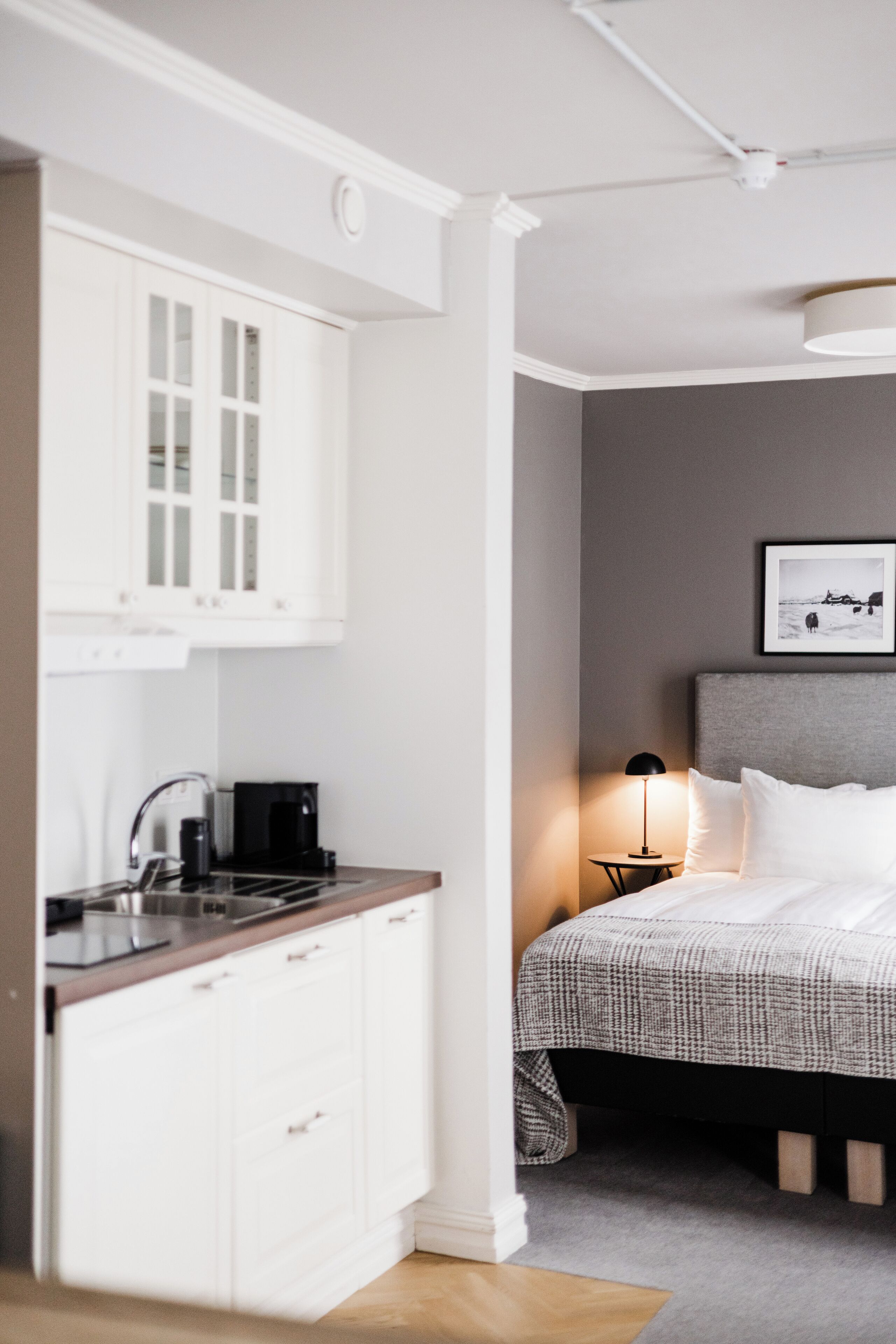 A modern hotel room with a kitchenette, a bed with a grey headboard, and a bedside table with a lamp.