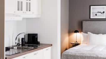 A modern hotel room with a kitchenette, a bed with a grey headboard, and a bedside table with a lamp.