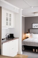 A modern hotel room with a kitchenette, a bed with a grey headboard, and a bedside table with a lamp.