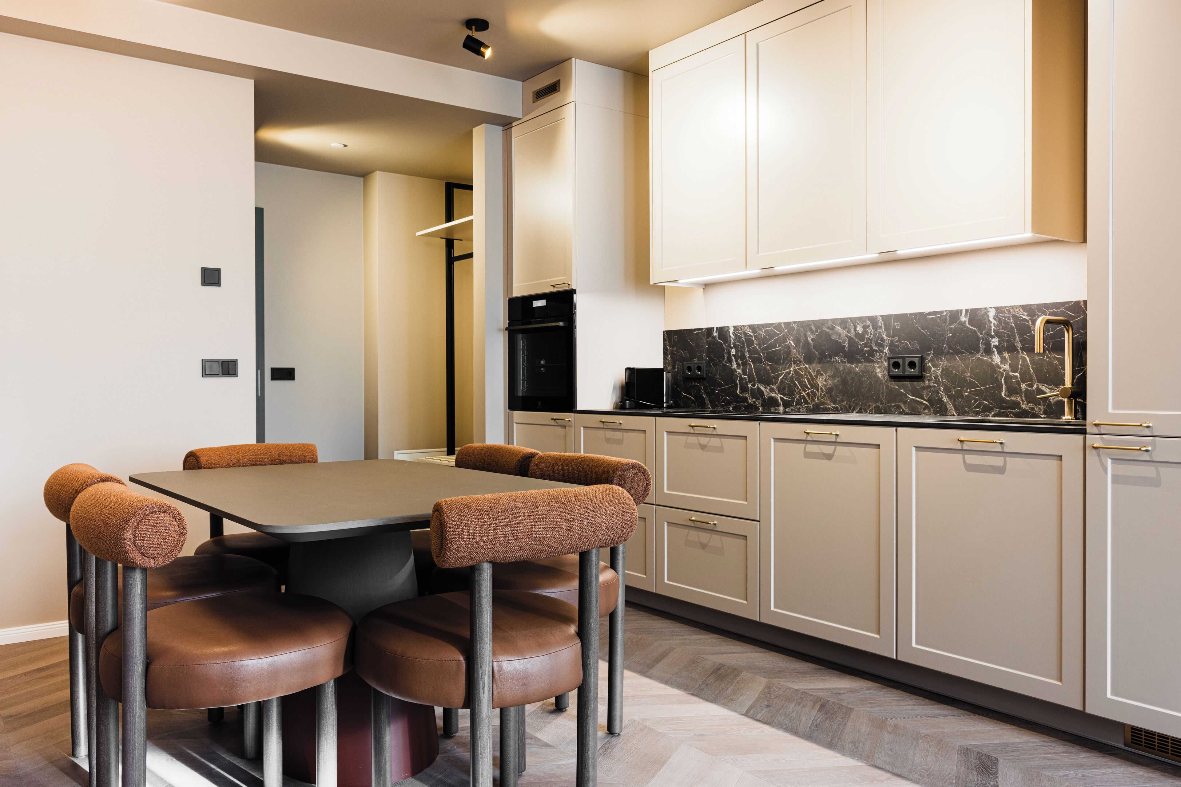 A modern kitchen with a marble countertop, beige cabinets, and a dining area with a dark table and brown chairs.
