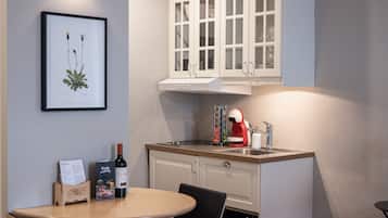 Una cocina compacta con armarios blancos, una mesa de comedor con dos sillas, una planta enmarcada en la pared y una botella de vino sobre la mesa.