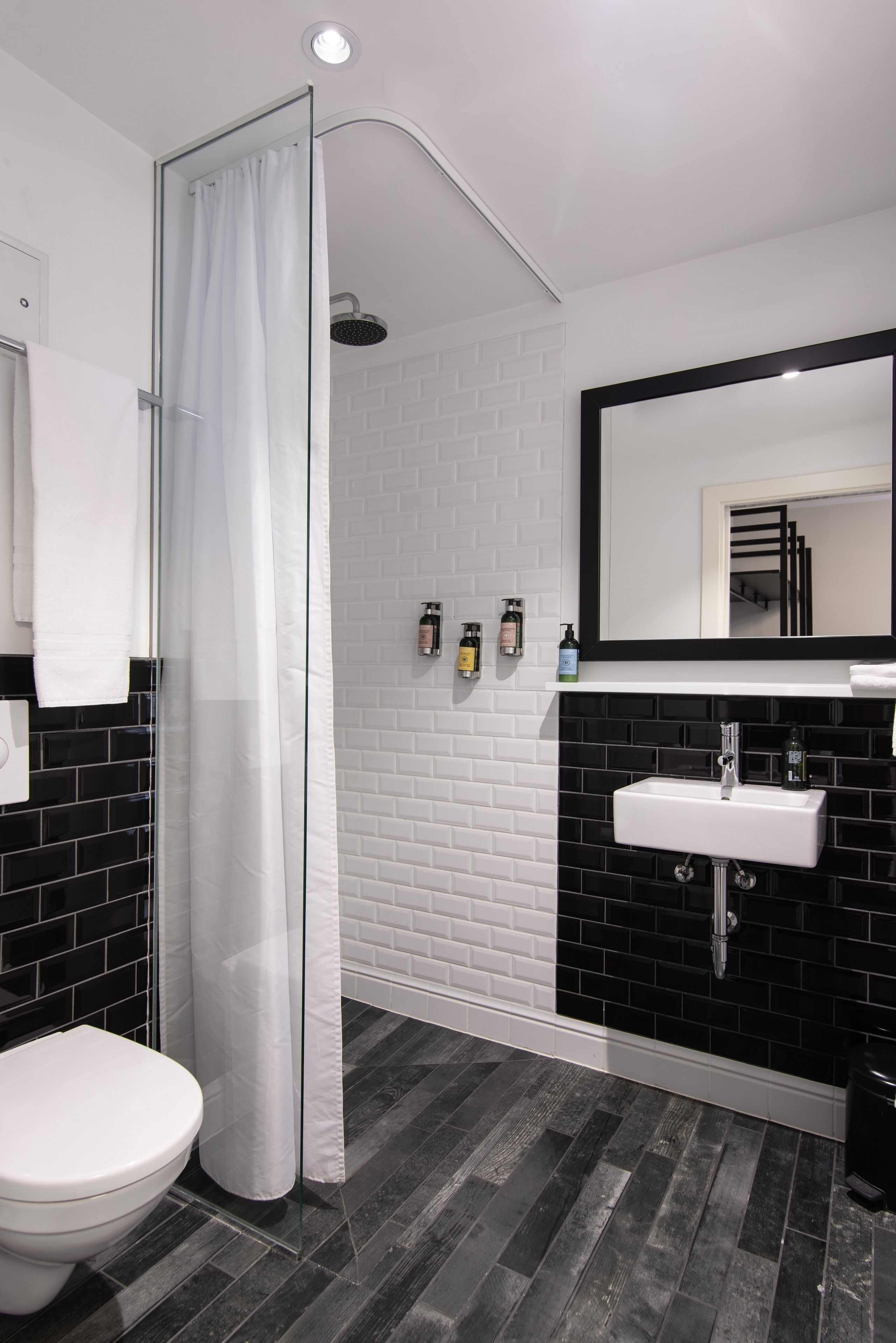 Een moderne badkamer met een douchecabine van glas, een wit toilet, een wand betegeld met zwart-wit tegels, een witte wastafel en een spiegel boven de wastafel.