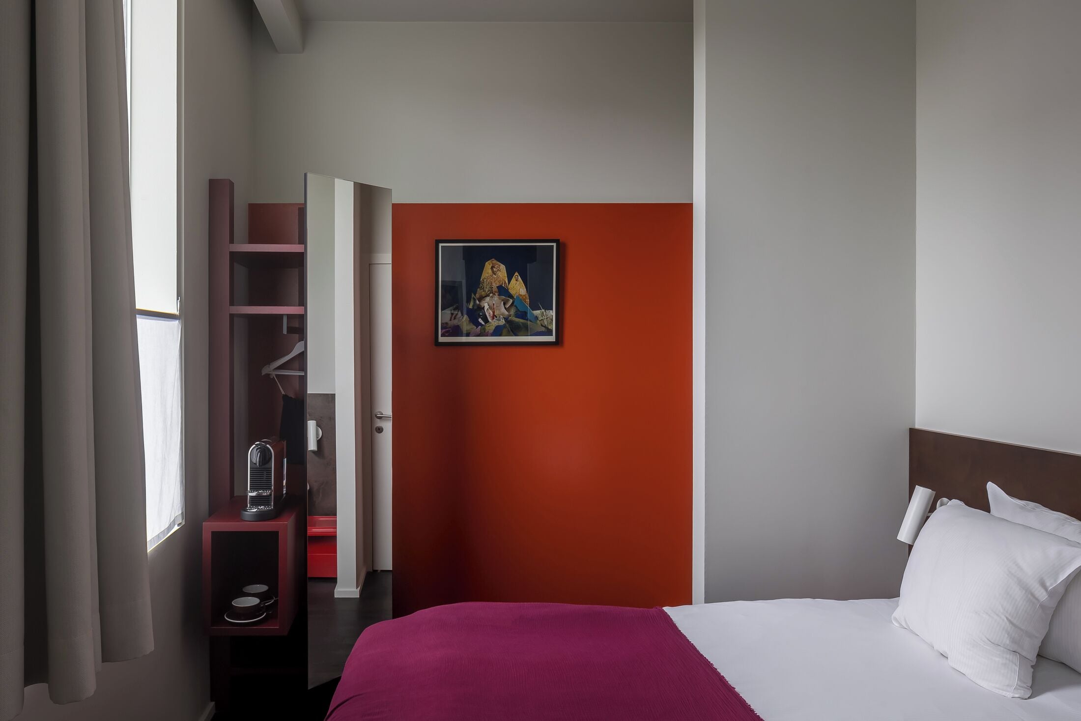 Een hotelkamer met een bed, een rode accentmuur, een schilderij en een nachtkastje met een lamp.