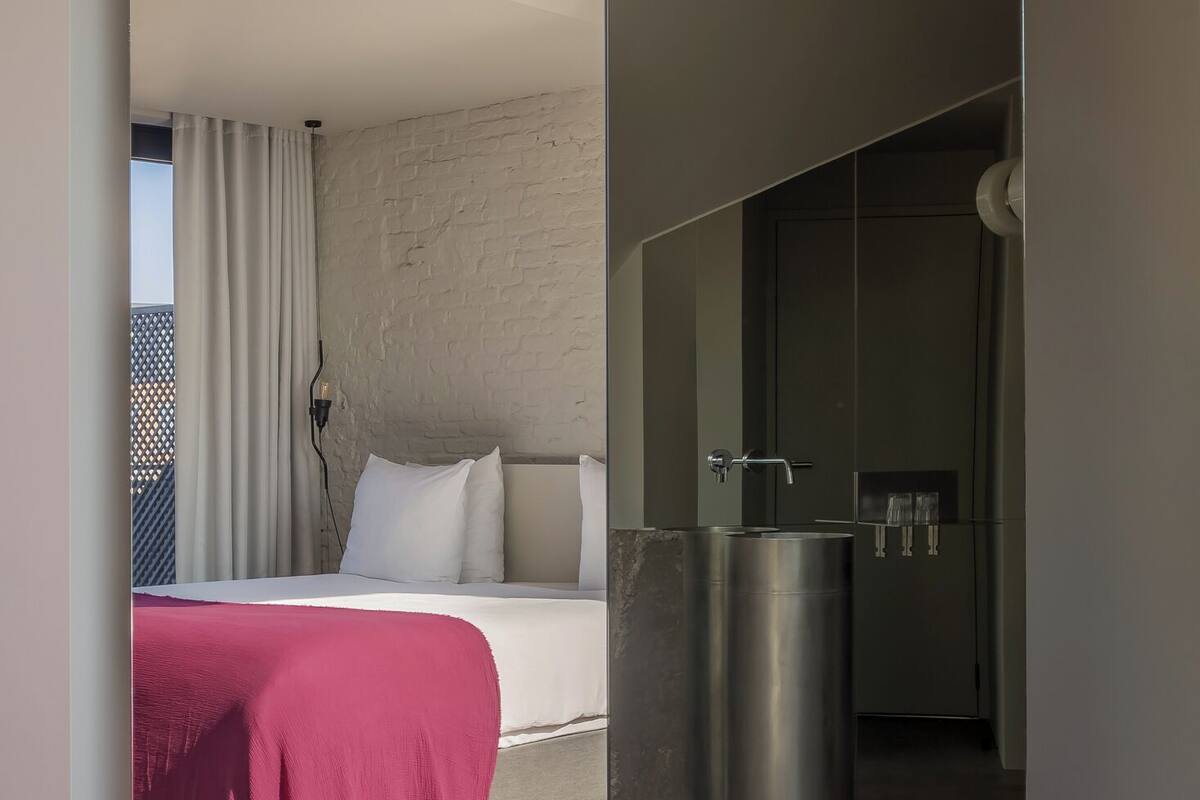 A modern hotel room with a large bed, a red throw blanket, and a small bathroom with a sink and mirror.