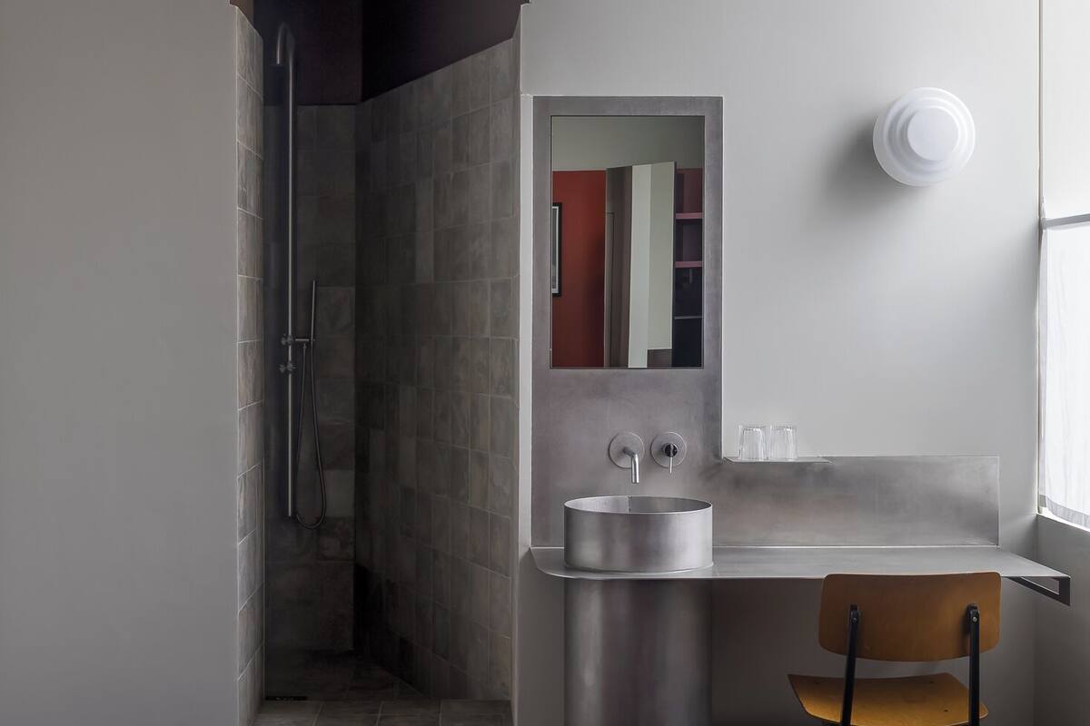 A modern hotel room with a bed, a desk with a chair, a bathroom with a shower, and a mirror.