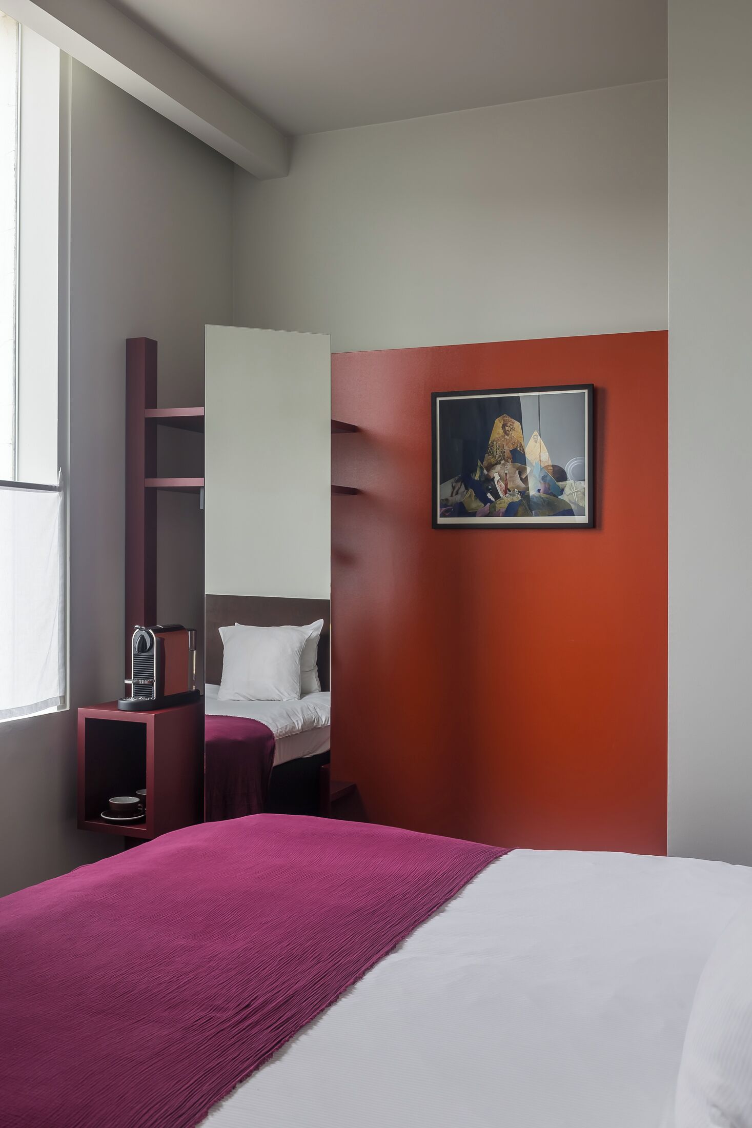 A modern hotel room with a bed, a small table with a coffee maker, a red accent wall, and a framed artwork.