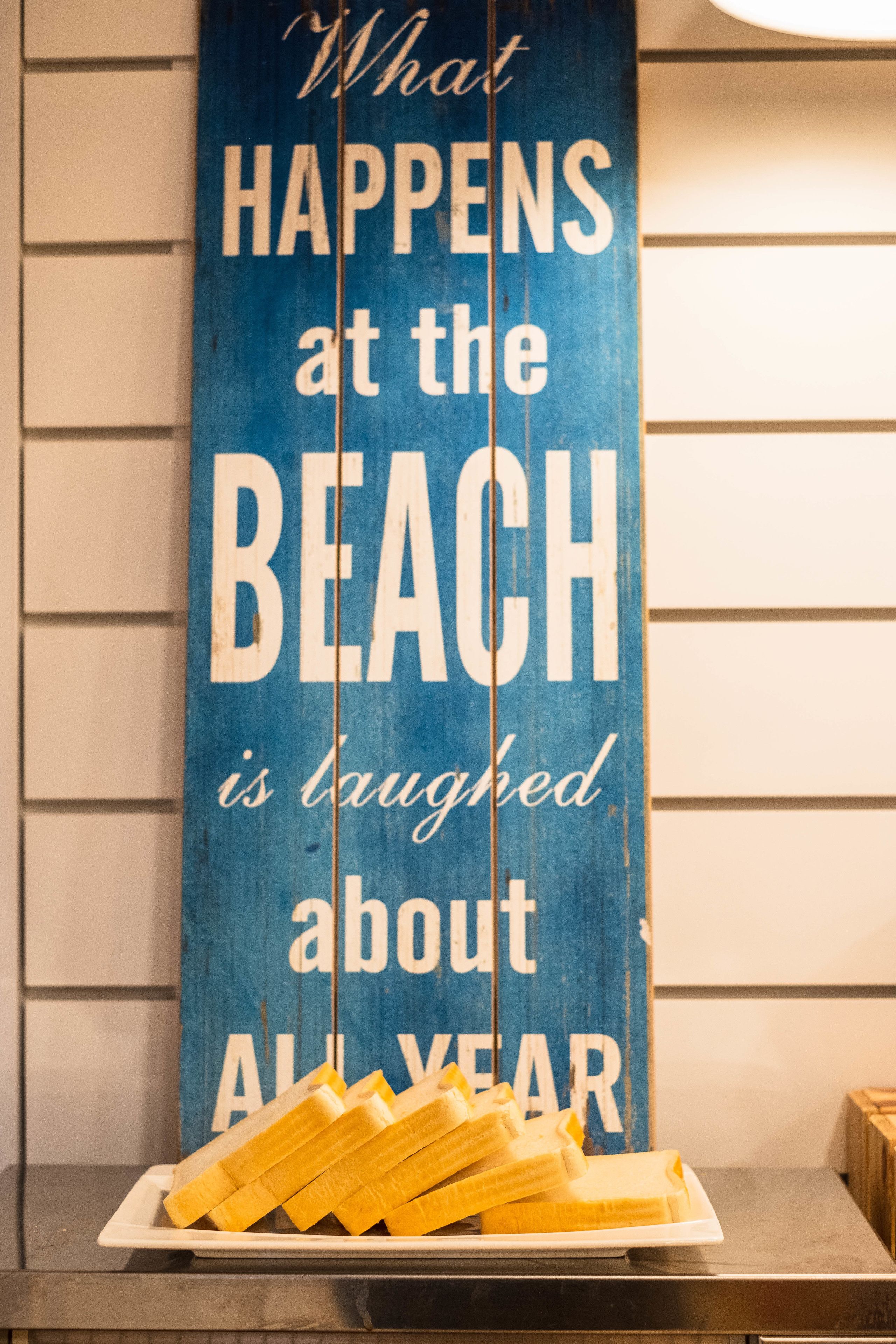 A blue sign with white text stating "What happens at the beach is laughed about all year" and a plate of bananas.