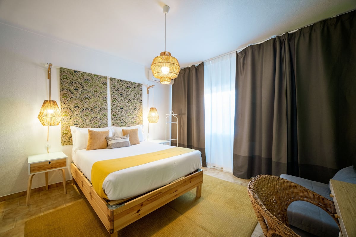 Een keurig opgeruimde hotelkamer met een groot bed, nachtlampjes, een rieten stoel en uitzicht op het omliggende landschap.