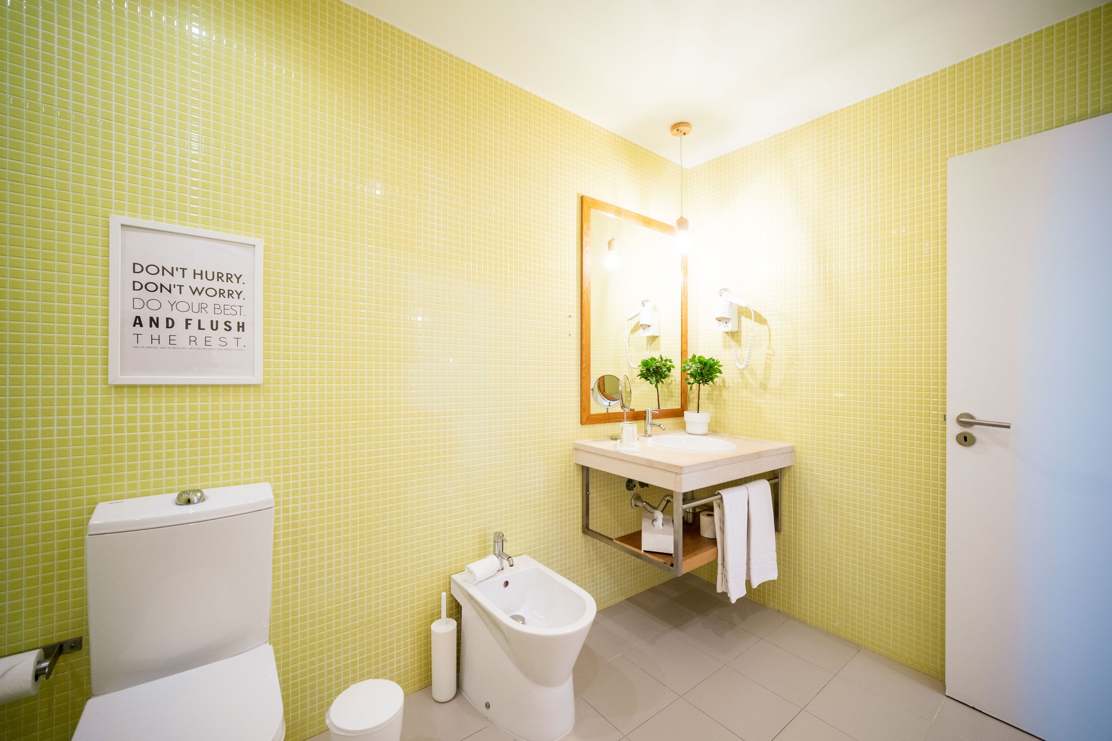 Een badkamer met geel betegelde wanden, een wit toilet, een bidet, een wastafel met spiegel, en een bord met een motiverende boodschap.