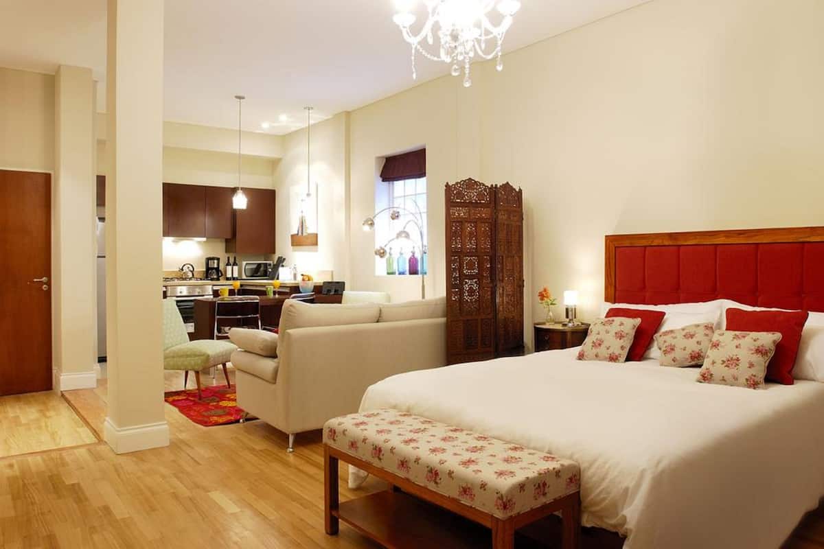 A hotel room with a large bed, a wooden headboard, a small bench, a kitchenette, a sofa, and a decorative wooden screen.