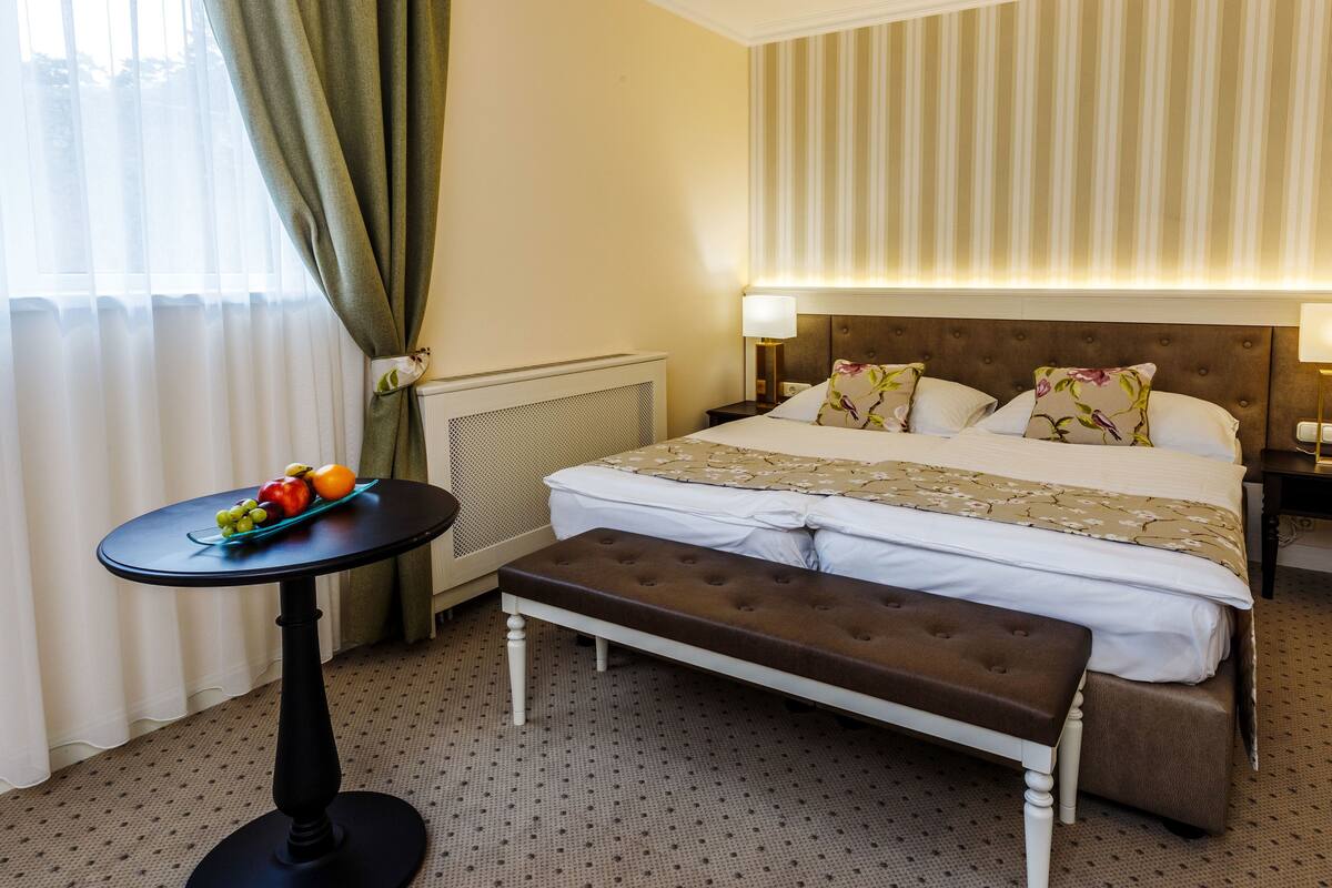 Une chambre d’hôtel avec un lit, des tables de chevet, un banc, une petite table avec des fruits et un radiateur.