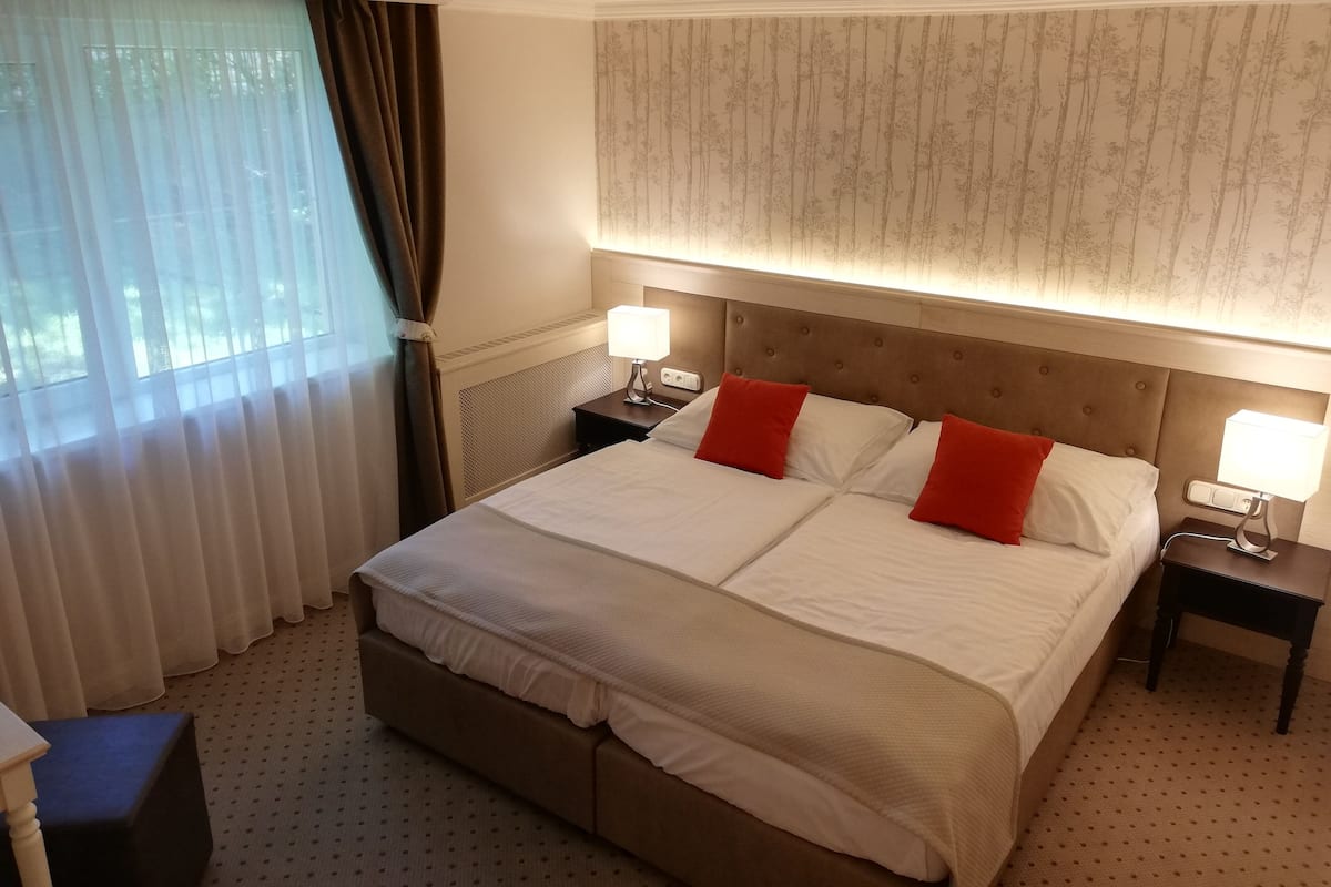 Une chambre d’hôtel avec un grand lit, deux tables de chevet avec des lampes, une fenêtre avec des rideaux et un papier peint à motifs sur le mur.