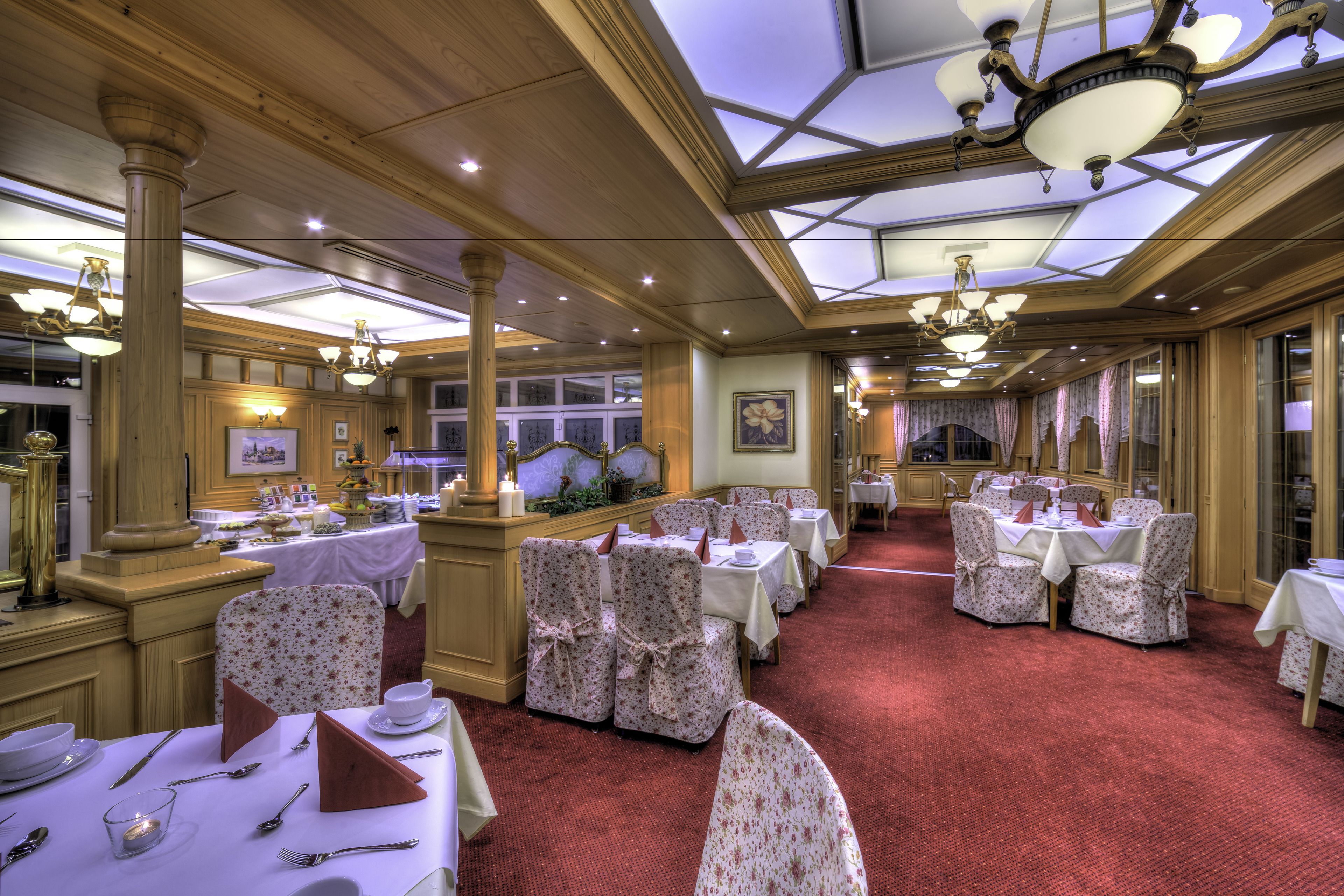 A formal dining area with multiple tables and chairs, a chandelier, and a red carpeted floor.