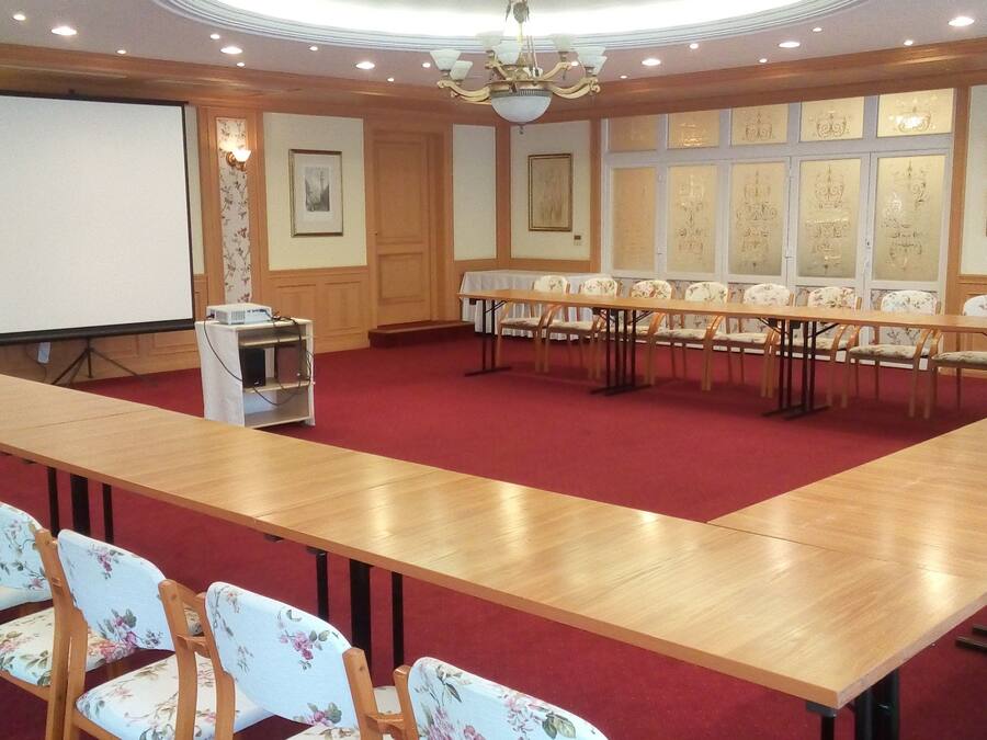 Un salón de conferencias con una larga mesa de madera, sillas tapizadas con motivos florales, una pantalla de proyector y cuadros en las paredes.