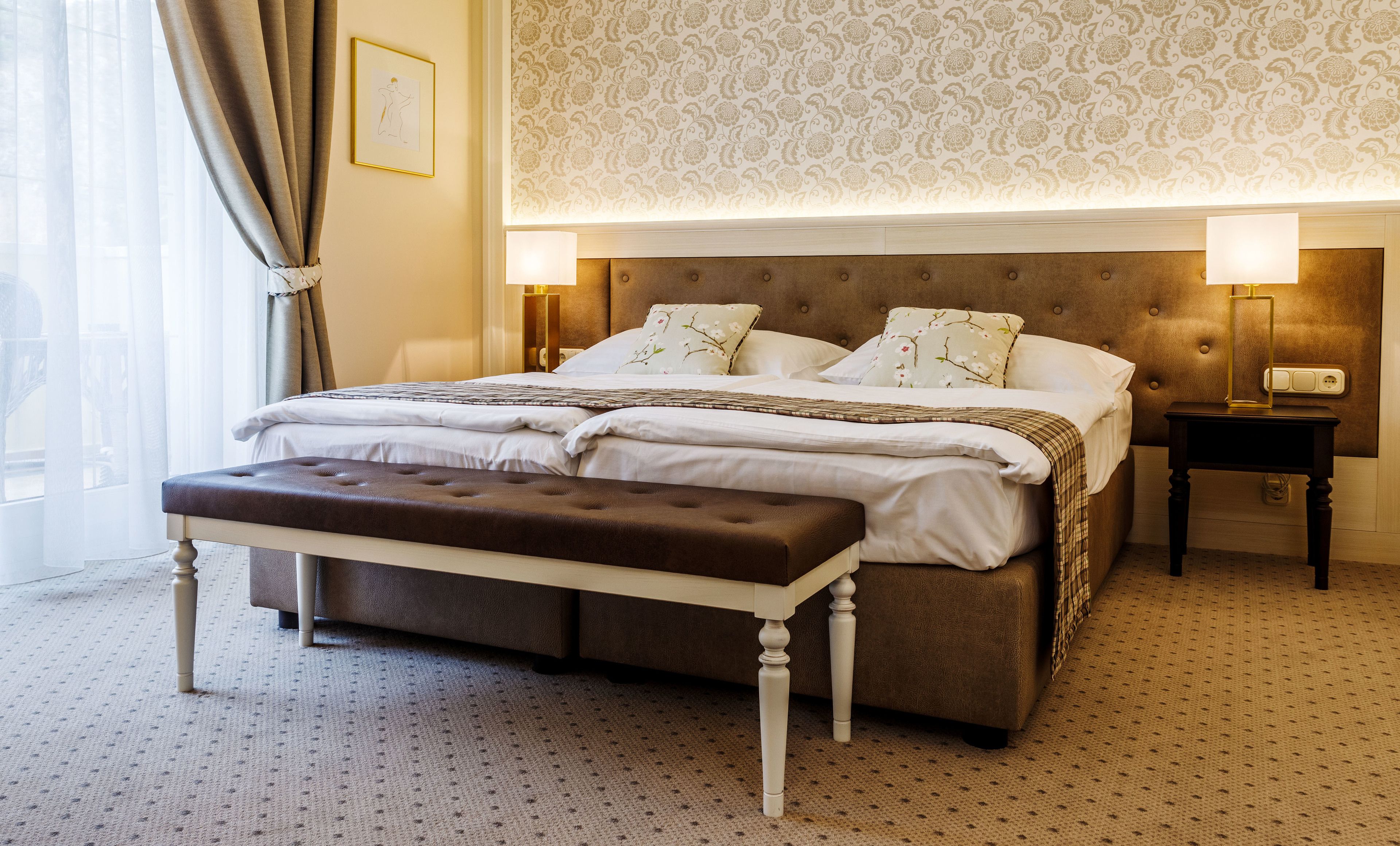 Un lit bien fait, avec une tête de lit marron, du linge de lit blanc et des coussins à motifs floraux. On y trouve un banc au pied du lit, une table de chevet avec une lampe et un luminaire fixé au mur.