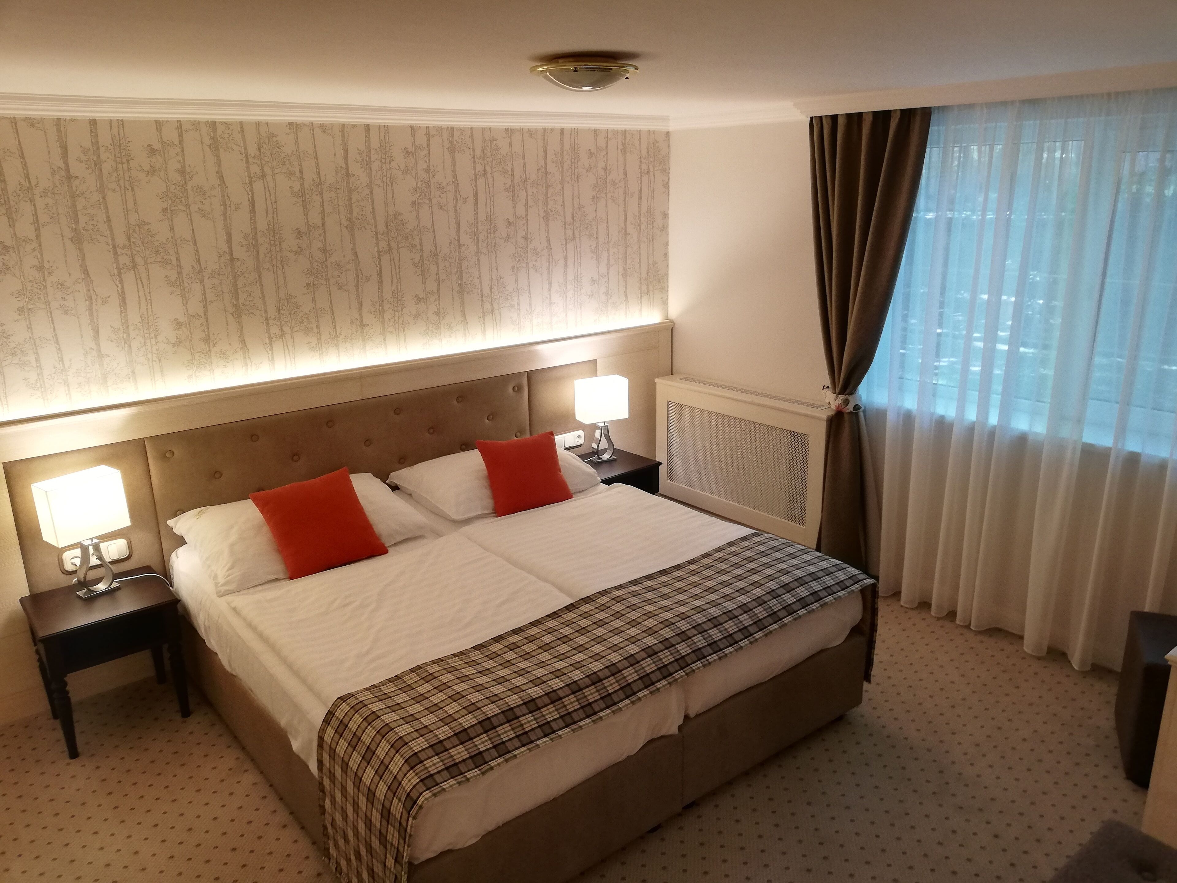 Habitación de hotel con una cama grande, dos mesitas de noche con lámparas, un radiador y una ventana con cortinas.