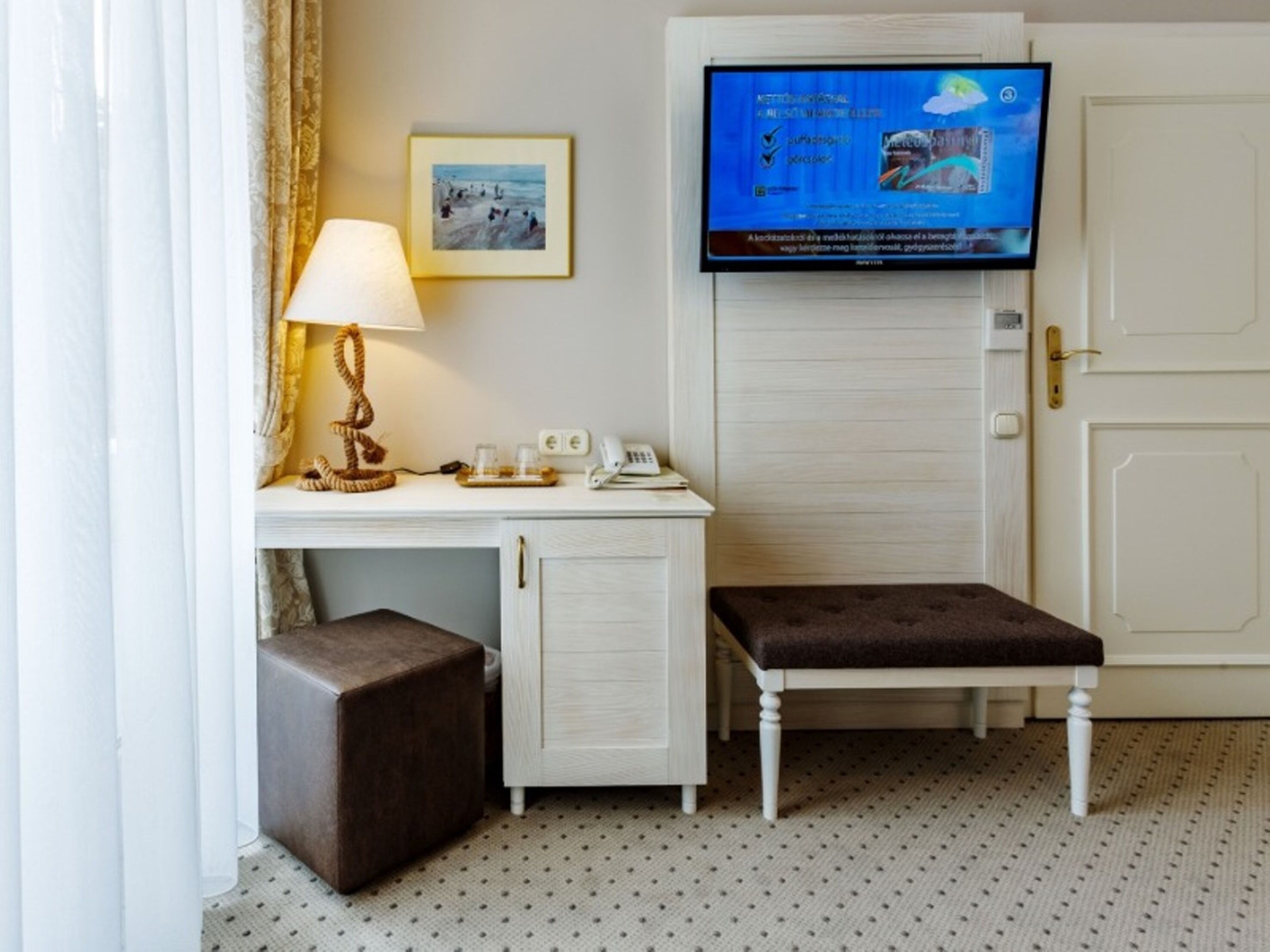 A hotel room with a desk, a bench, a television, and a framed picture on the wall.