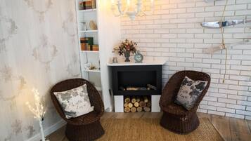 A cozy room with a fireplace, two wicker chairs with cushions, a small shelf with books and decorative items, and a wall-mounted knife rack.