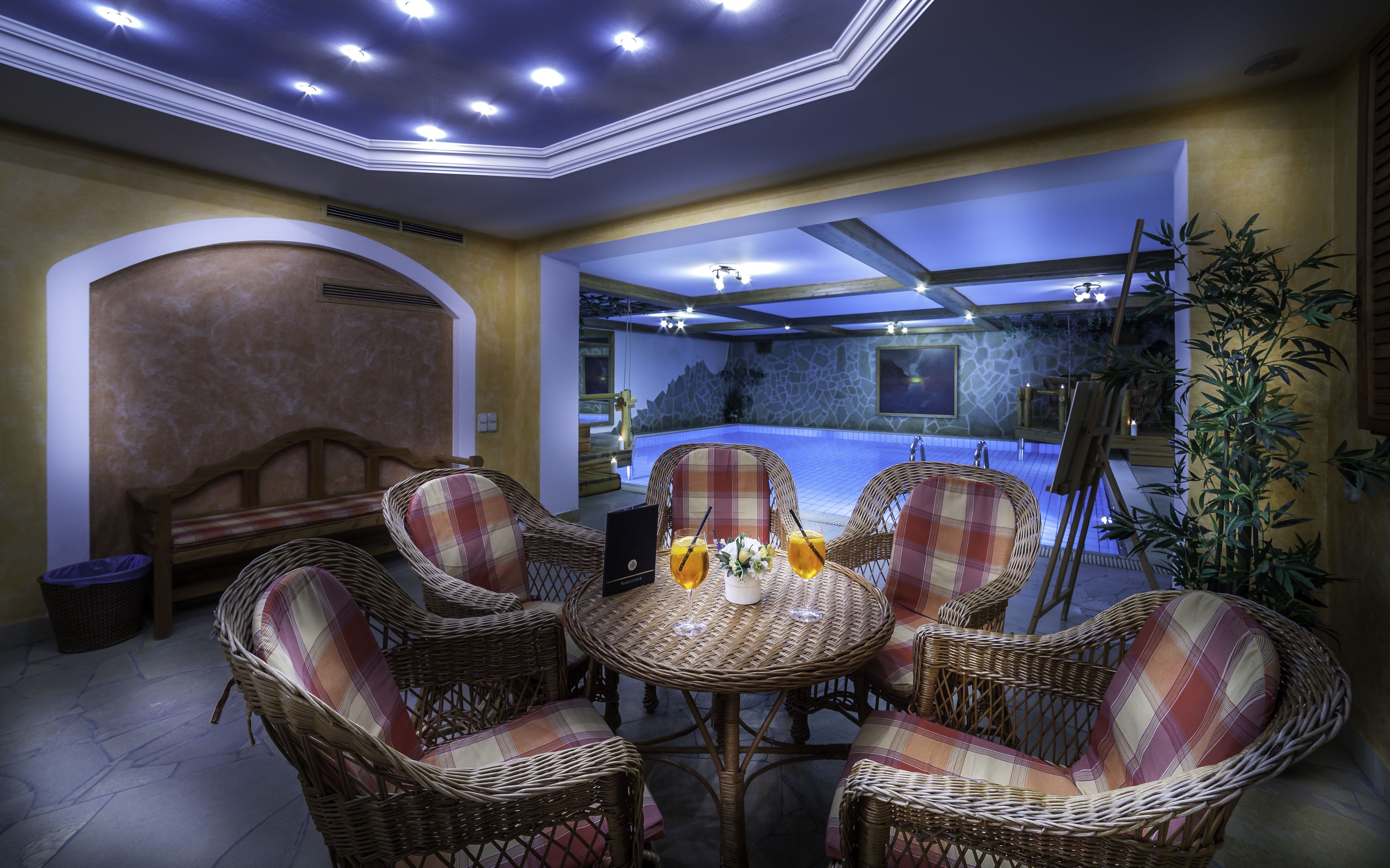 Un comedor con sillas de mimbre y una mesa, una piscina al fondo y un techo con iluminación empotrada.