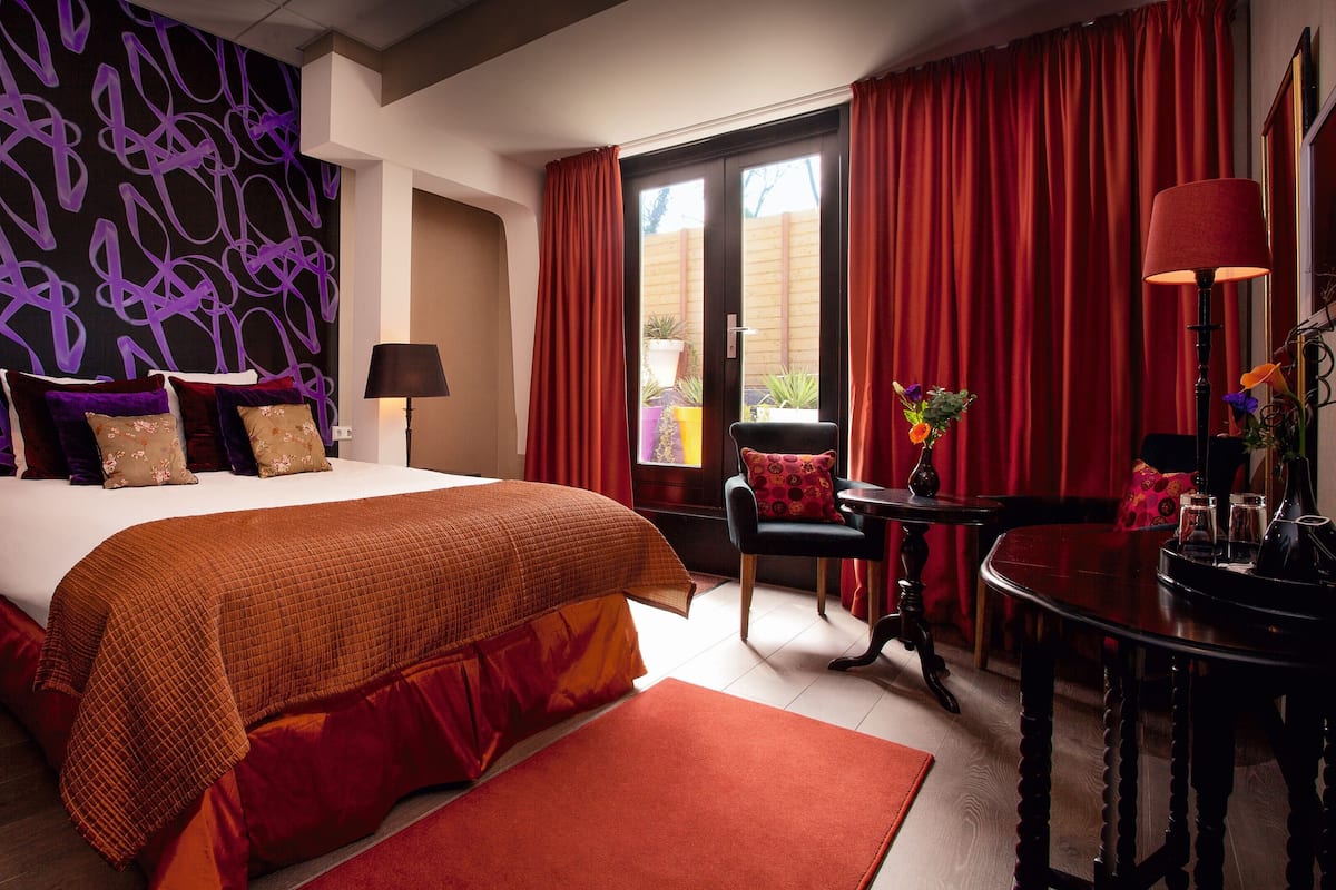 A hotel room with a large bed, red curtains, a small table with a vase of flowers, and a chair.