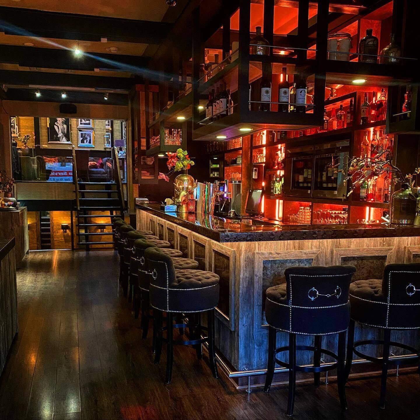 Zona de bar con varios taburetes, una barra llena de botellas y una escalera que sube a un nivel superior.