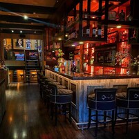 Zona de bar con varios taburetes, una barra llena de botellas y una escalera que sube a un nivel superior.
