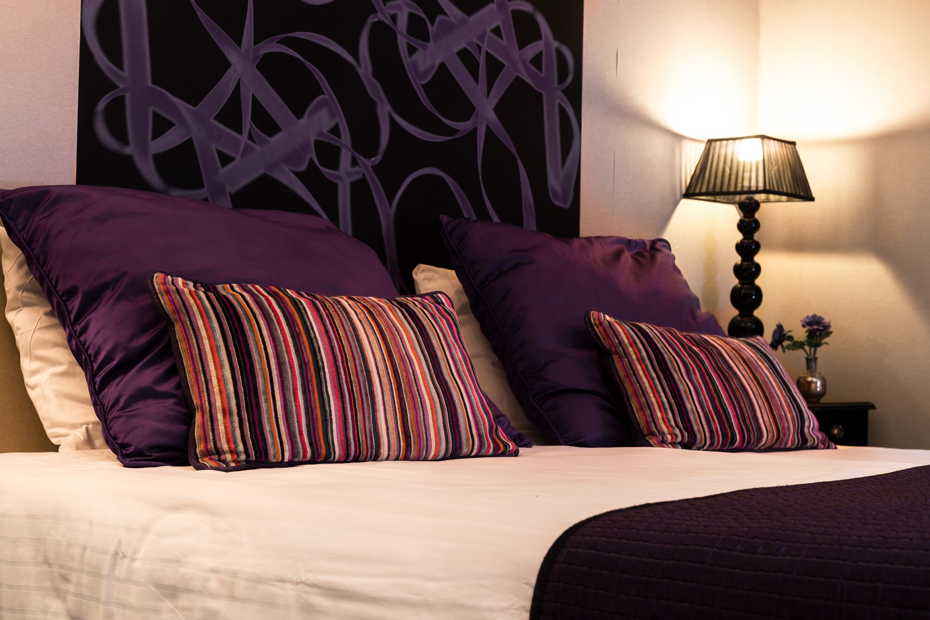 A bed with two large purple pillows and a striped pillow, a bedside lamp, and a wall with a geometric design.