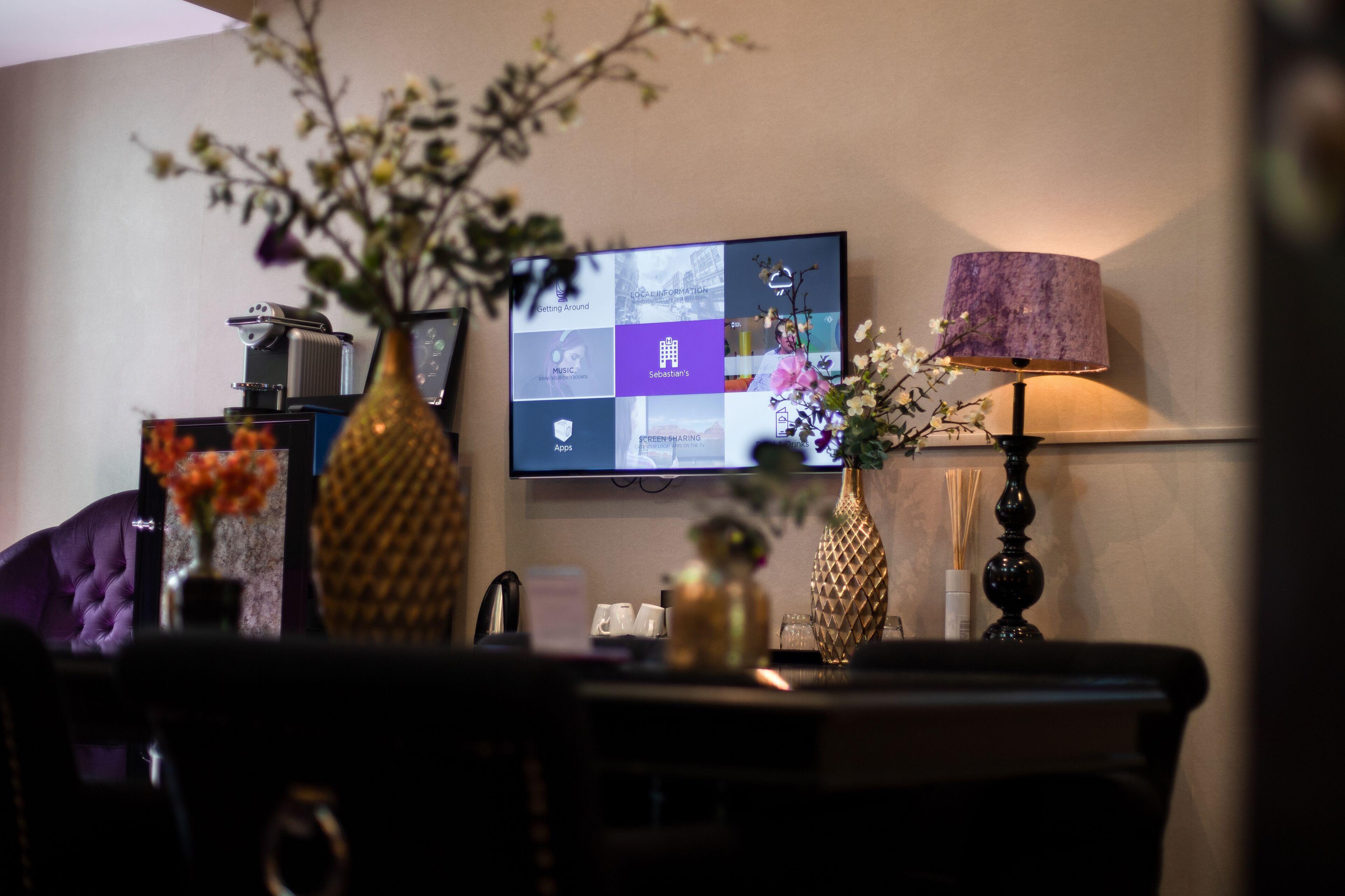 A modern living room with a flat-screen TV mounted on the wall, a decorative vase with flowers, a lamp, and a coffee machine on a table.