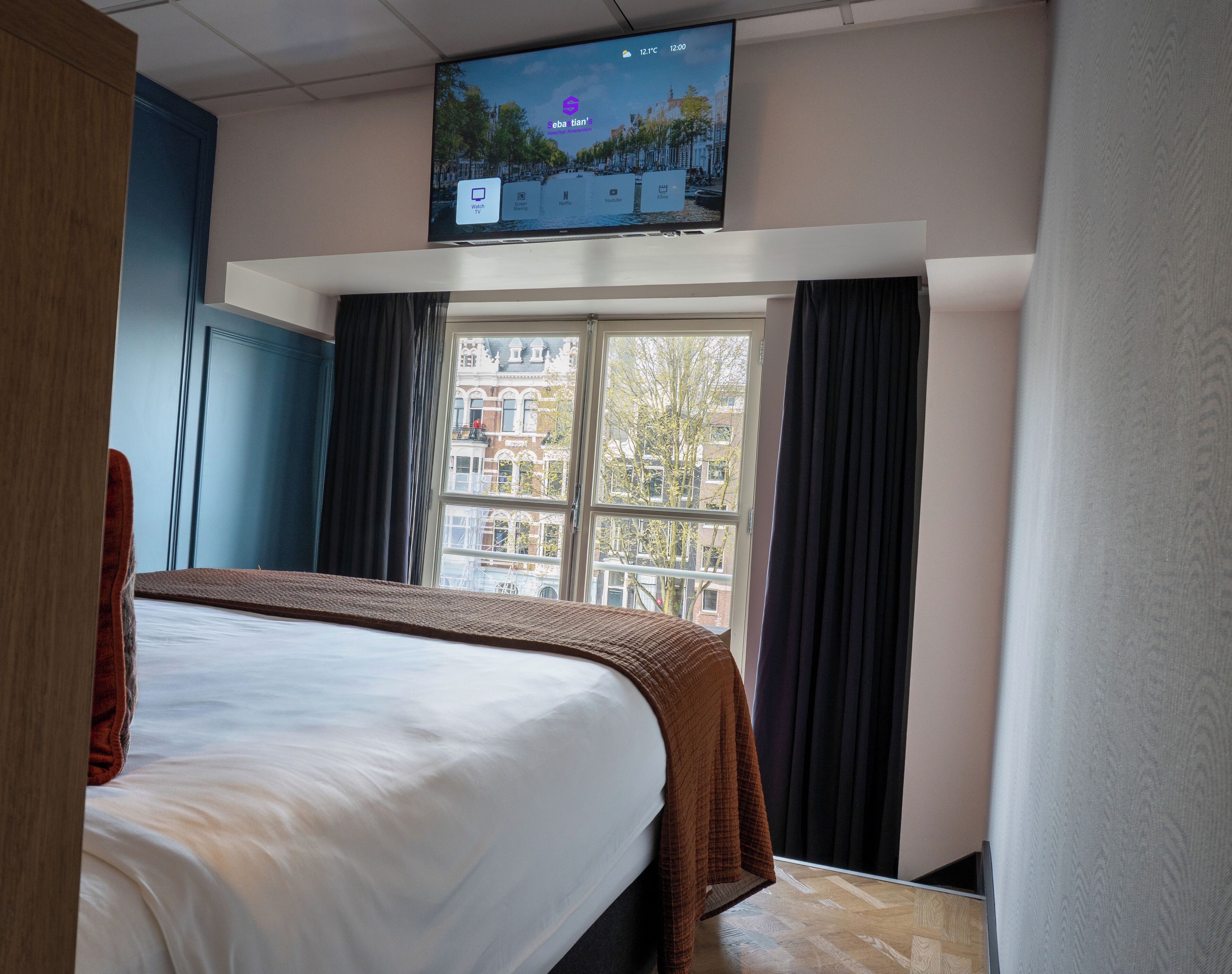 A hotel room with a bed, a TV mounted on the wall, a window with curtains, and a view of buildings outside.