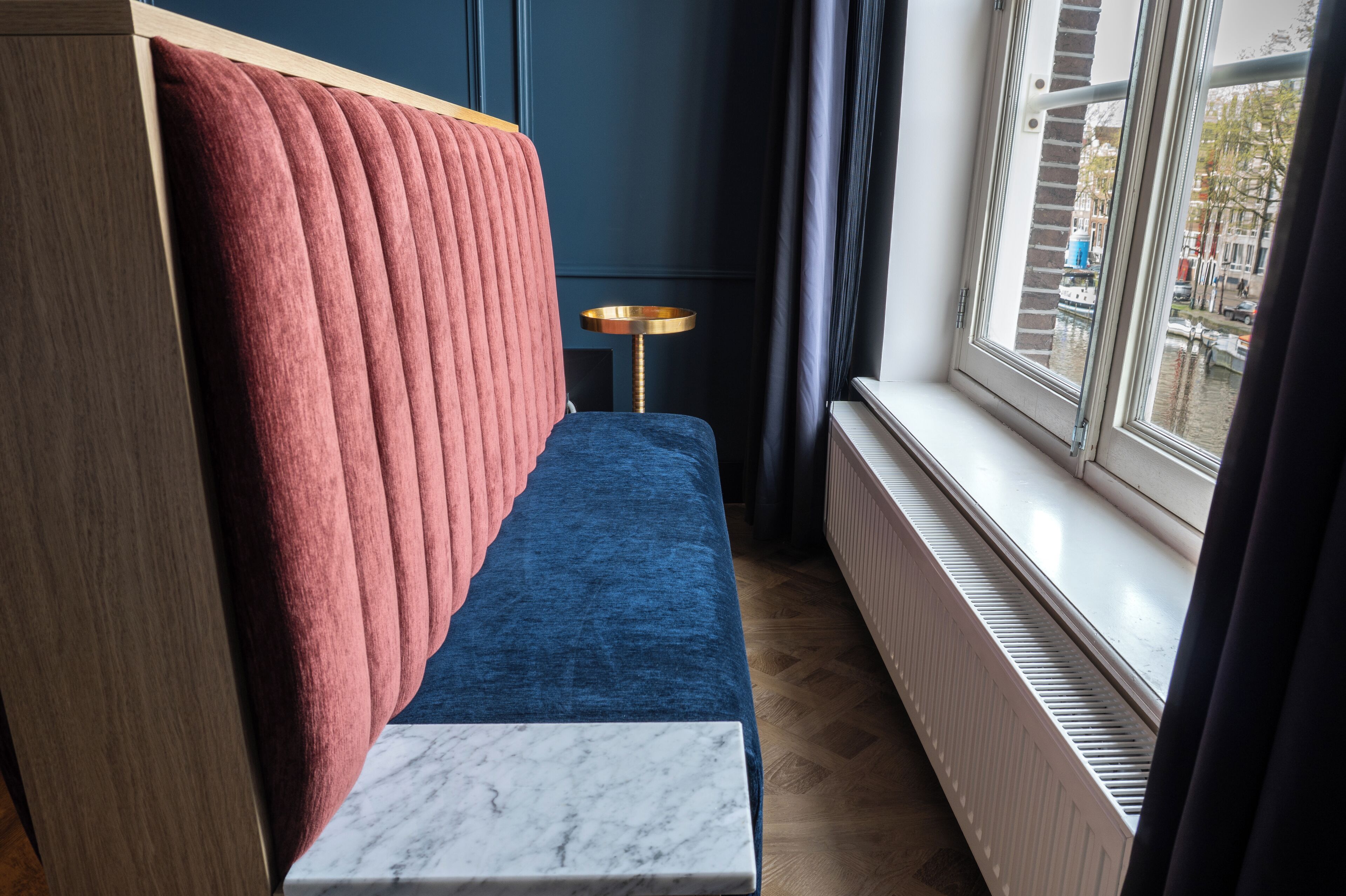A room with a blue sofa, a red upholstered headboard, a small round table, and a window with a view of the city.