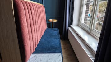 Une pièce avec un canapé bleu, une tête de lit rouge en tissu, une petite table ronde et une fenêtre donnant sur la ville.