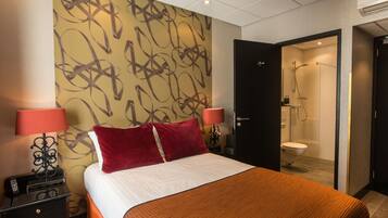 Habitación de hotel con cama, dos lámparas de noche, teléfono, mesita de noche, zona de ducha y un inodoro.