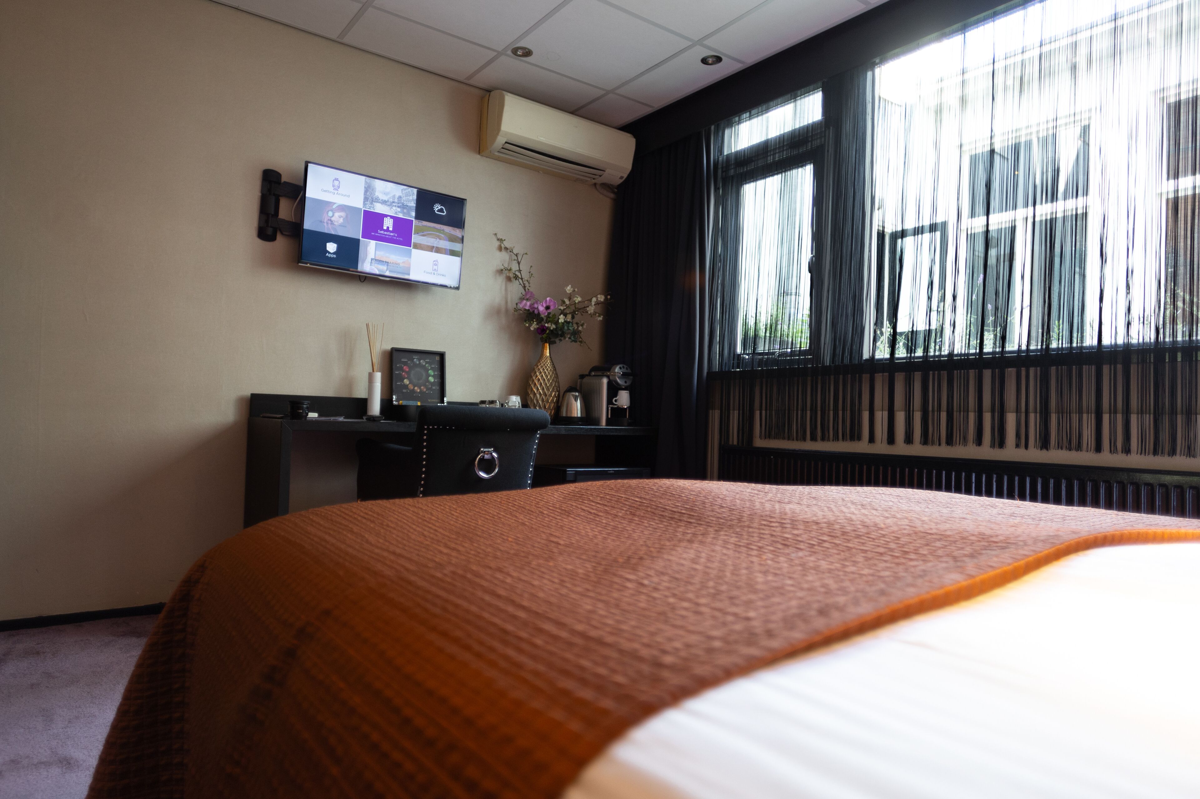 A hotel room with a bed, a desk with a computer monitor, a vase with flowers, and a window with curtains.