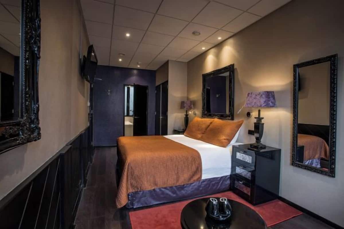 A hotel room with a bed, bedside table, lamp, mirror, and a red rug.