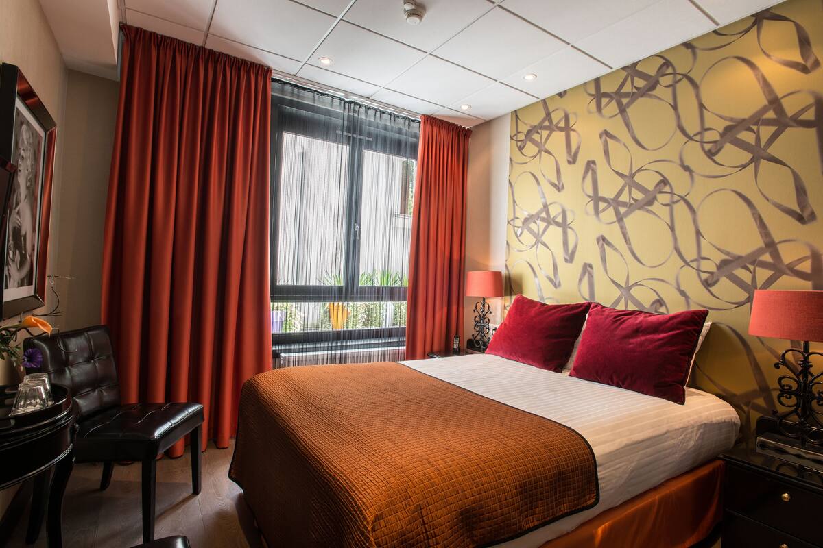 A hotel room with a large bed, red curtains, a desk with a chair, and a wall with abstract patterns.
