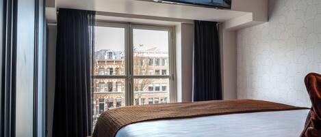 A hotel room with a bed, a television mounted on the wall, a window with curtains, and a patterned wall.