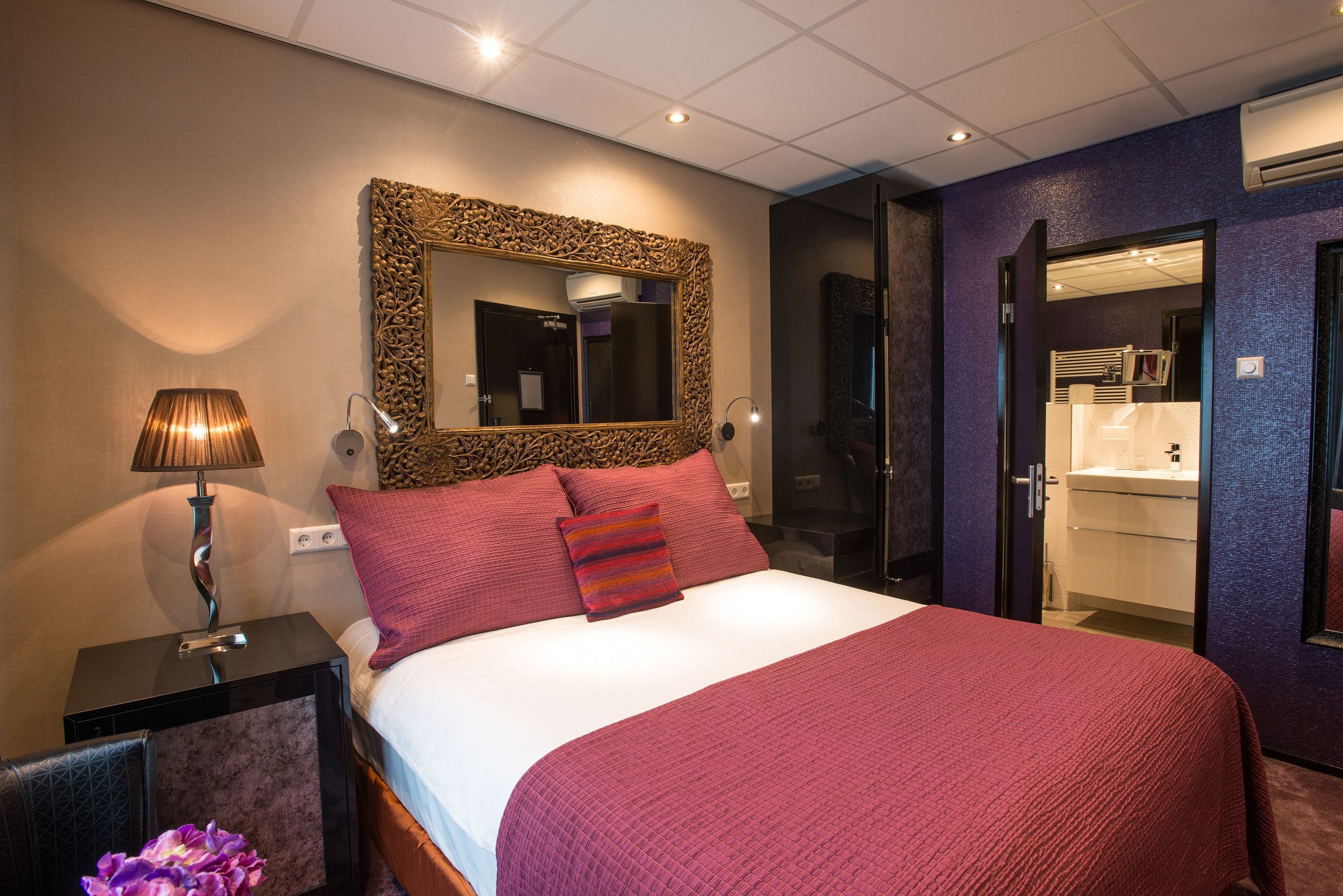 Habitación de hotel con una cama grande, un espejo con marco decorativo, una lámpara de noche y vista al baño.