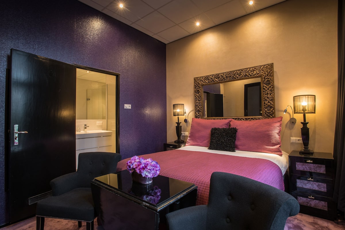 A hotel room with a bed, two chairs, a table with a vase, a mirror, bedside lamps, and a wardrobe.