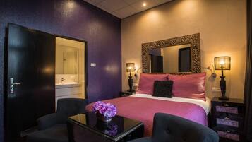 A hotel room with a bed, two chairs, a table with a vase, a mirror, bedside lamps, and a wardrobe.