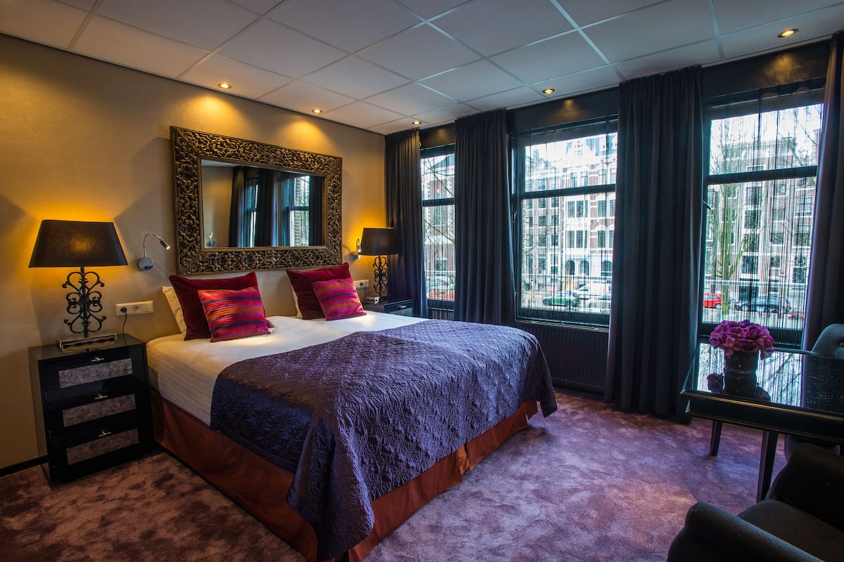 A hotel room with a large bed, two bedside tables, a mirror, and a view of the city through large windows.